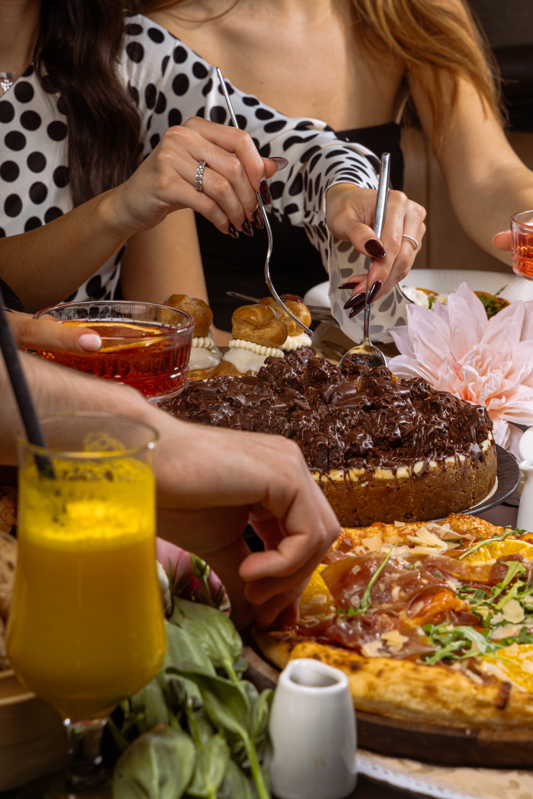 People sharing chocolate cake, pizza, and drinks at a lively dinner table