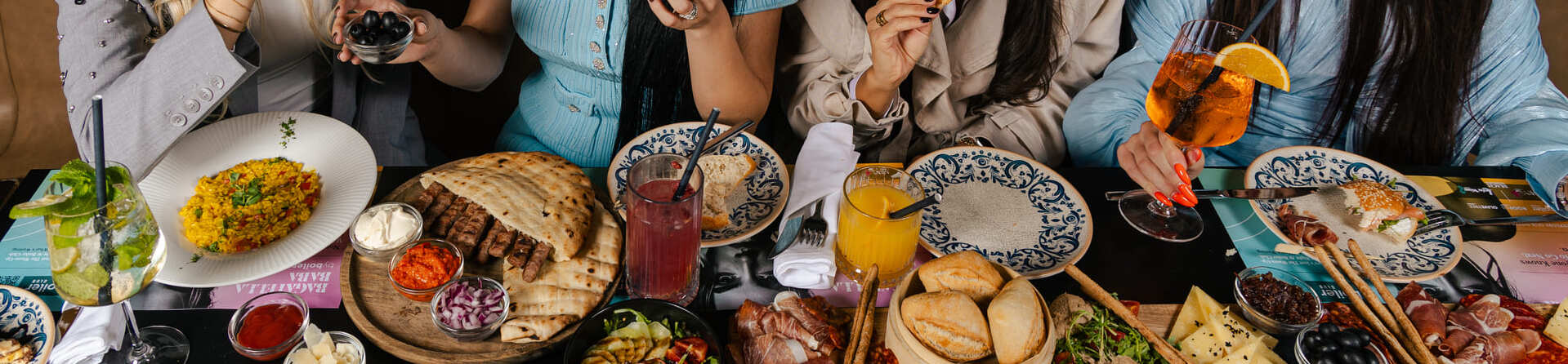 Four women seated at a restaurant table enjoying a variety of Mediterranean dishes and drinks, smiling and sharing food in a lively social setting.