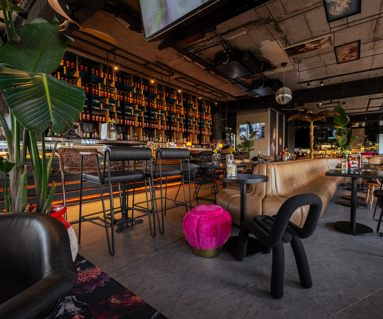 Modern upscale bar with industrial-style decor, featuring a long leather banquette, high-top tables, vibrant green plants, a wall of backlit liquor bottles, and eclectic furniture including a bright pink stool and black sculptural chair.