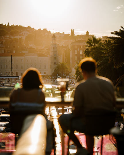 Two people enjoy cocktails at a rooftop bar overlooking a historic European city at sunset, with church towers, palm trees, and harbor in the distance.