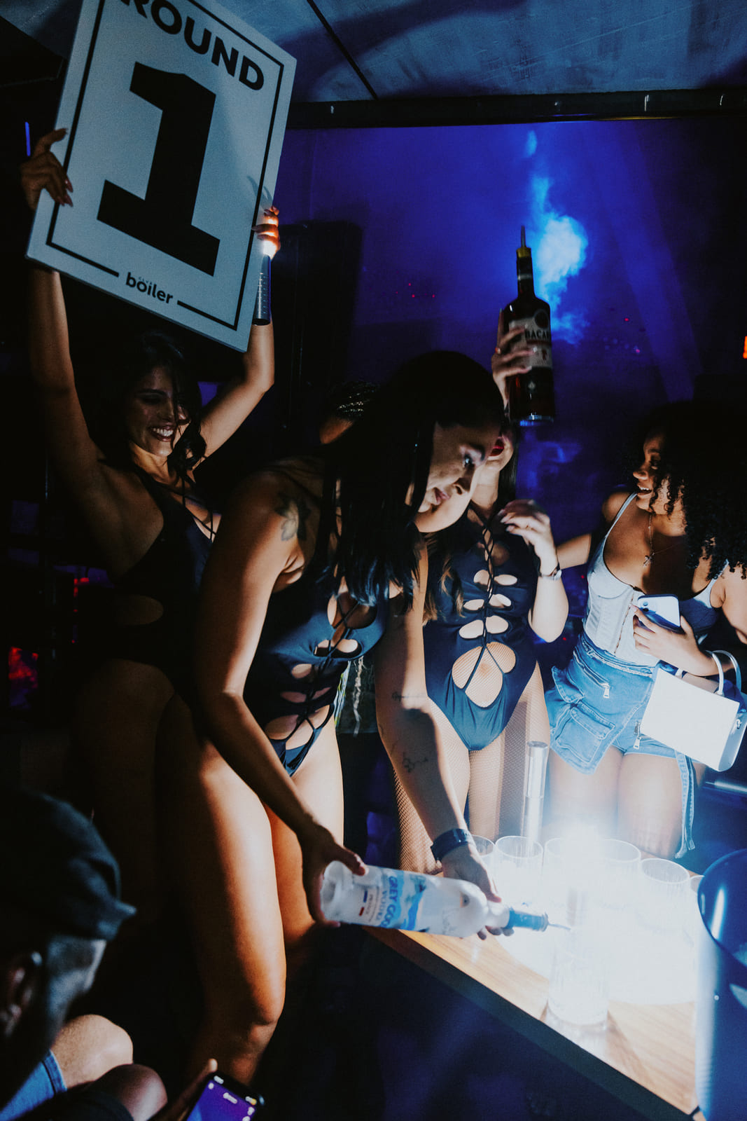 Women in black cutout bodysuits pouring drinks for guests at a nightclub, one holding a “Round 1” sign.