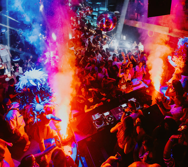 Crowded nightclub with vibrant lighting, smoke effects, and dancers in feathered costumes performing near the DJ booth.