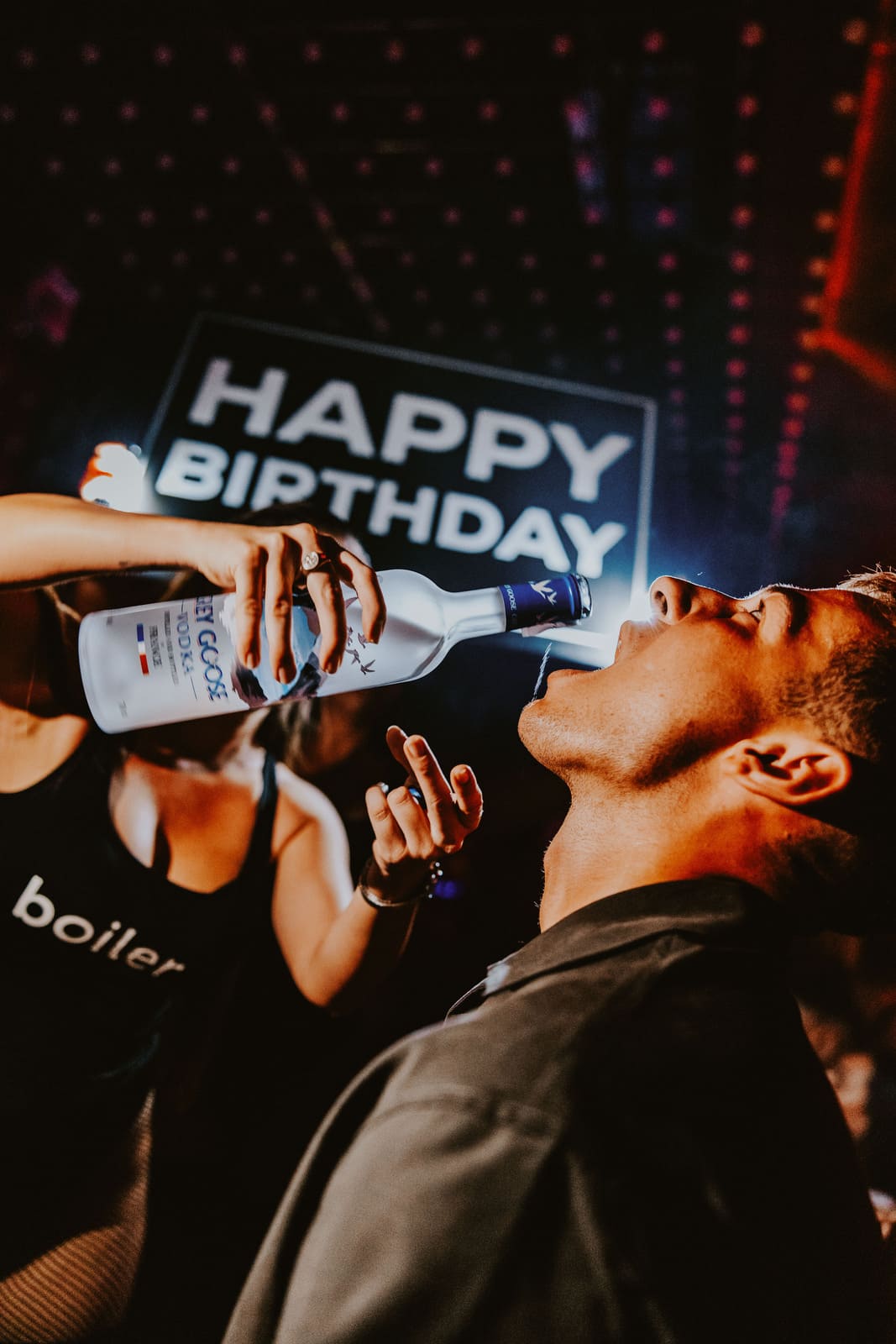 Club host pouring Grey Goose directly into a guest’s mouth under a “Happy Birthday” sign.