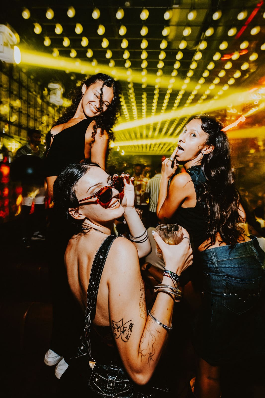 Three women posing with drinks and smiling under yellow club lights on a crowded dance floor.