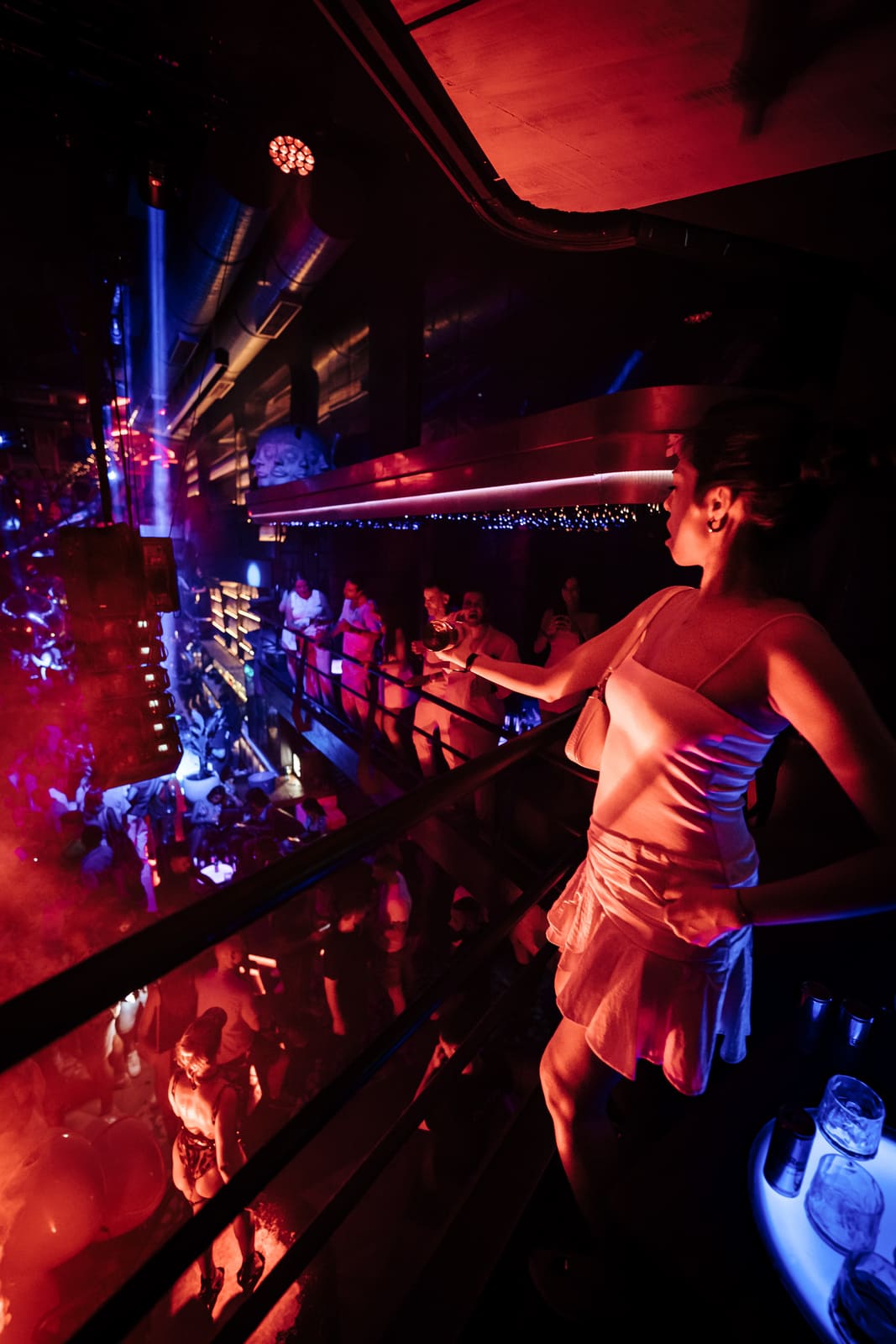 Woman in white dress enjoying the view from the upper level of a packed nightclub with red lighting.