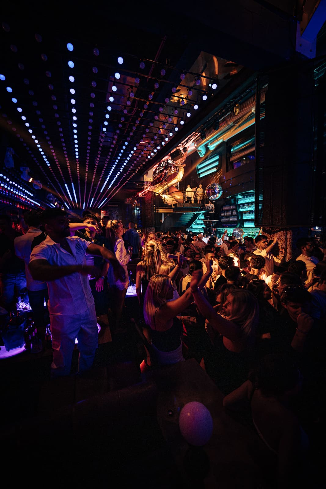 Wide view of a lively nightclub crowd under LED ceiling lights and neon signs.