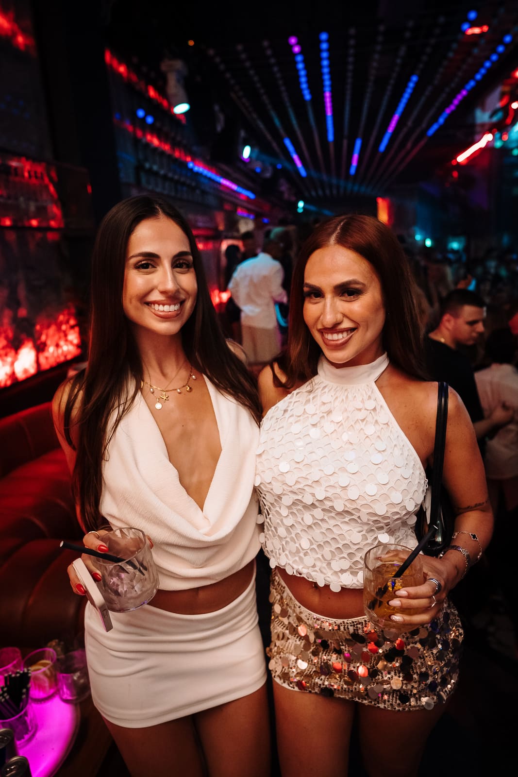 Two women dressed in white smiling and holding drinks in a vibrant club setting.