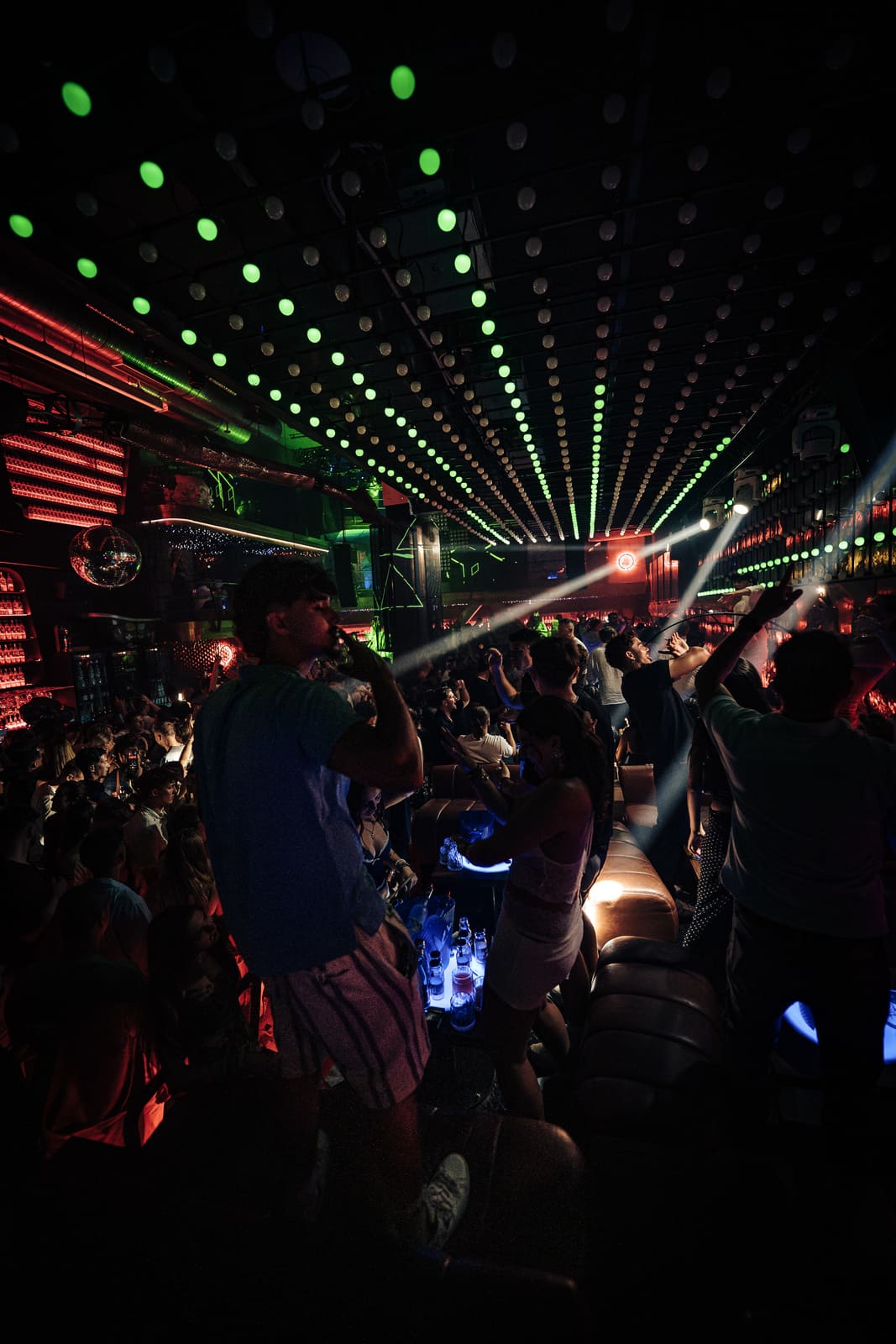 Clubgoers dance on couches under a ceiling of green lights and lasers.