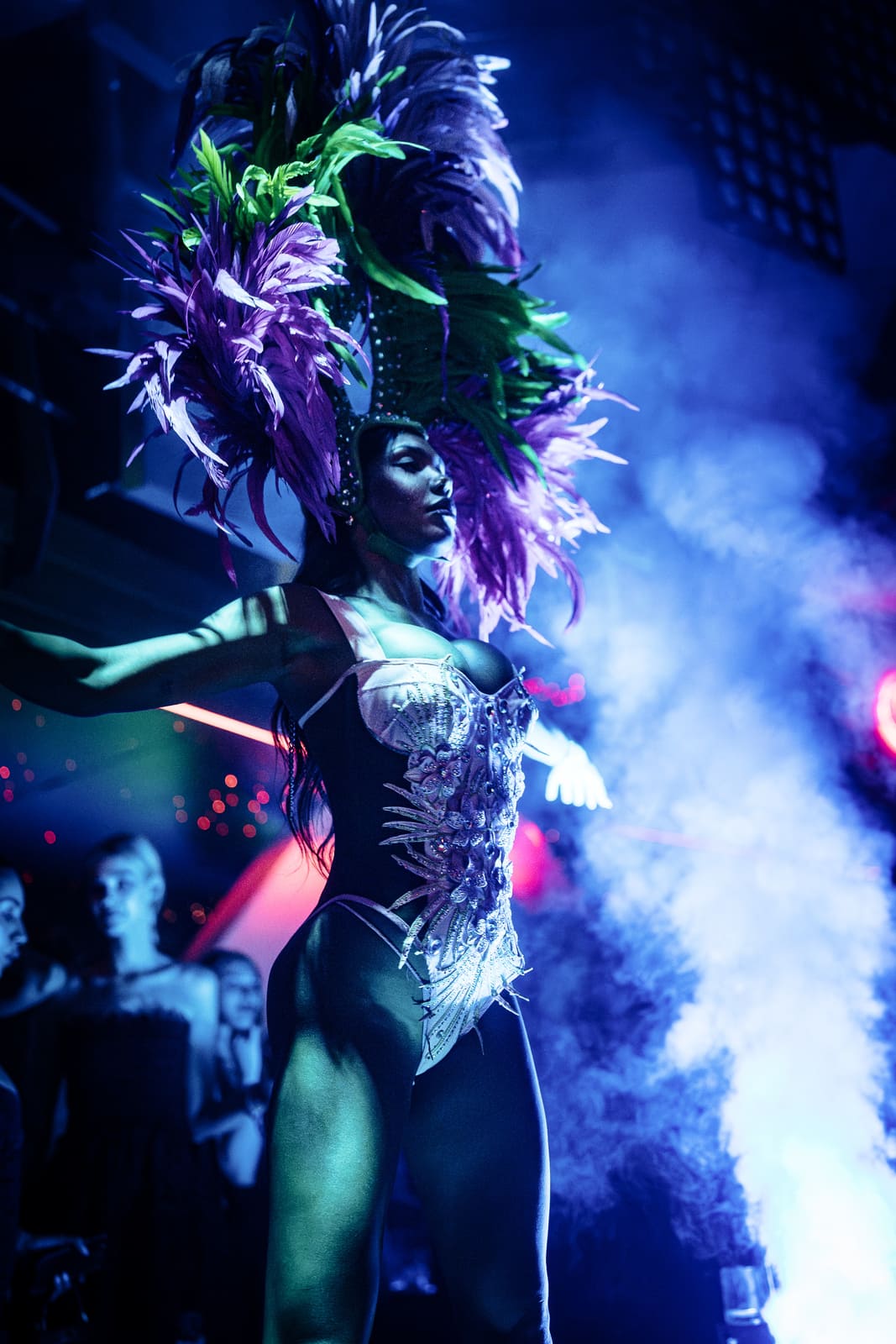 Female performer in ornate bodysuit and tall purple and green feathered headpiece posing under blue stage lights.