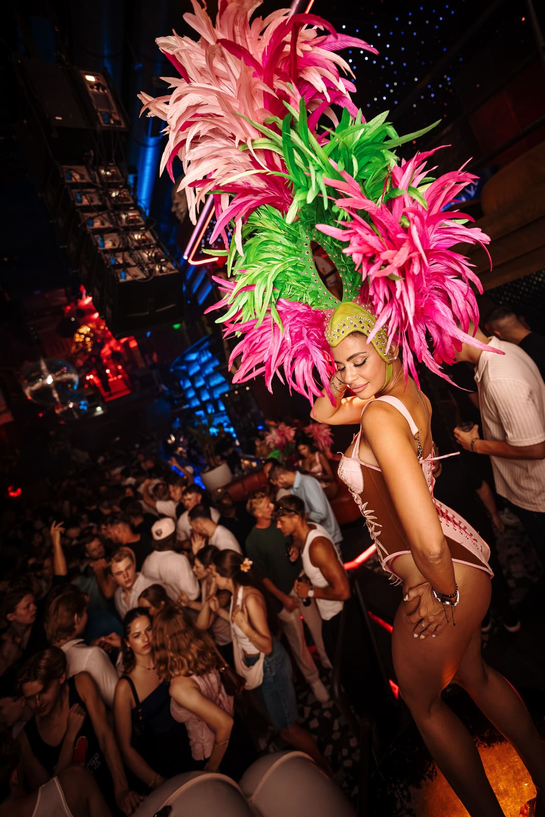 Smiling dancer in a pink and green feathered costume standing above a crowded nightclub dance floor.