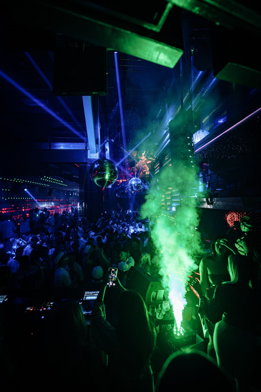Nightclub crowd dancing under green fog, disco balls, and laser lights with DJ booth in foreground.
