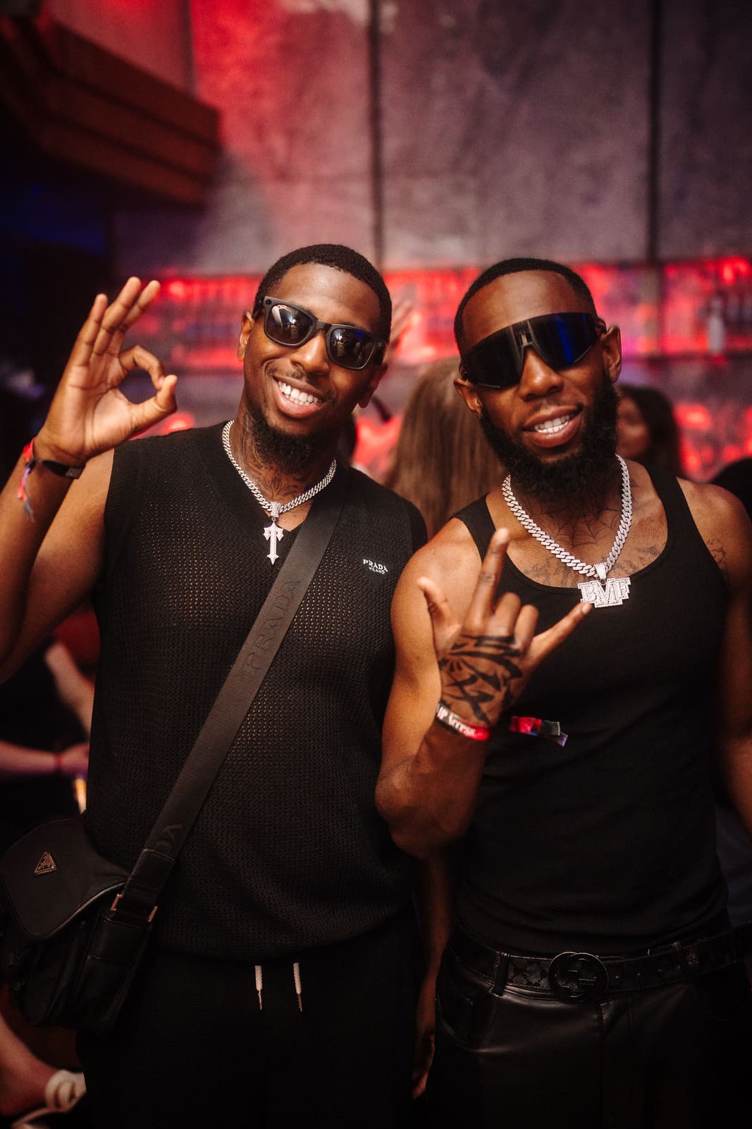 Two men in black outfits pose and smile under red nightclub lighting.