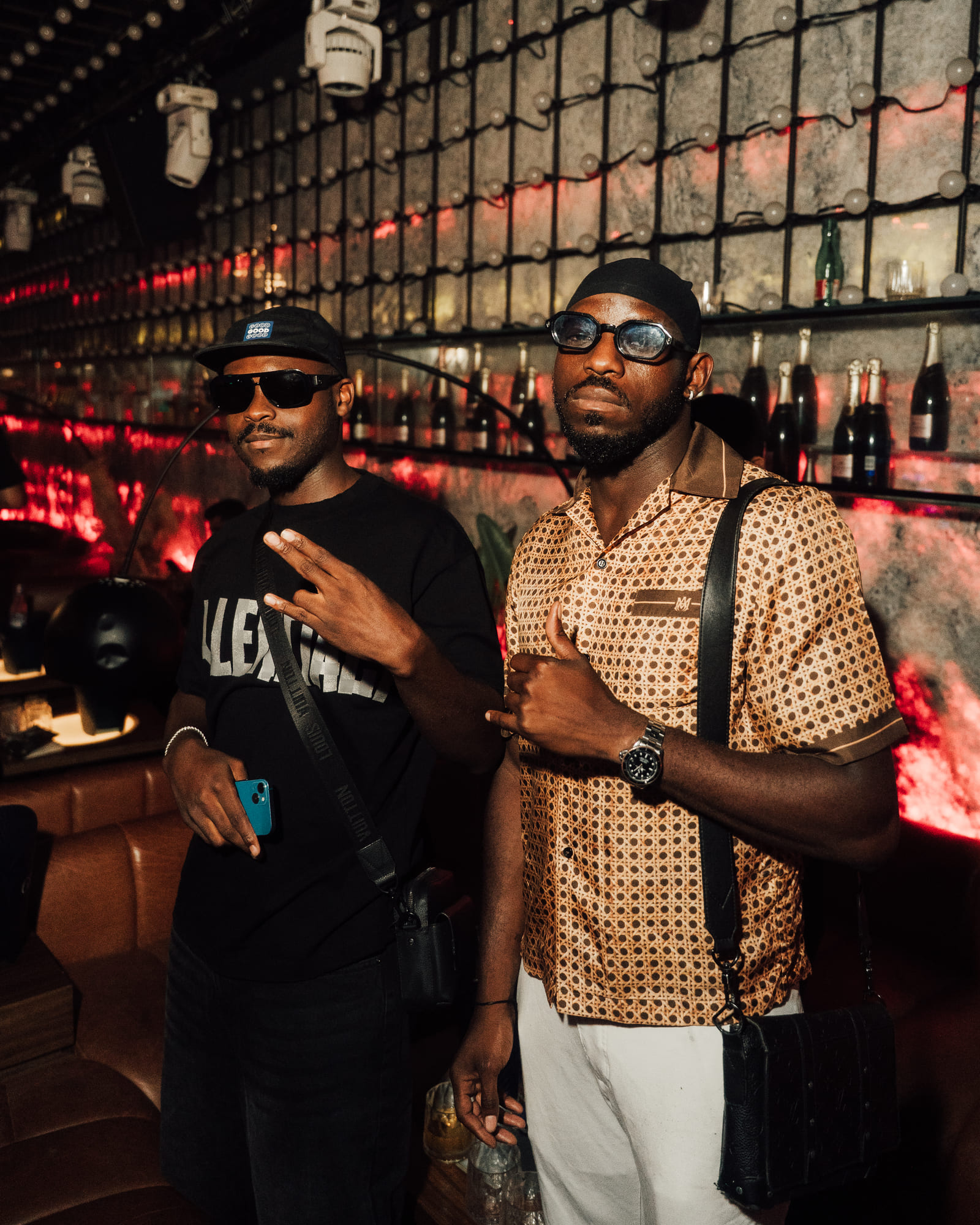 Two stylish men standing and posing inside a nightclub lounge area, with champagne bottles displayed in the background and red ambient lighting.