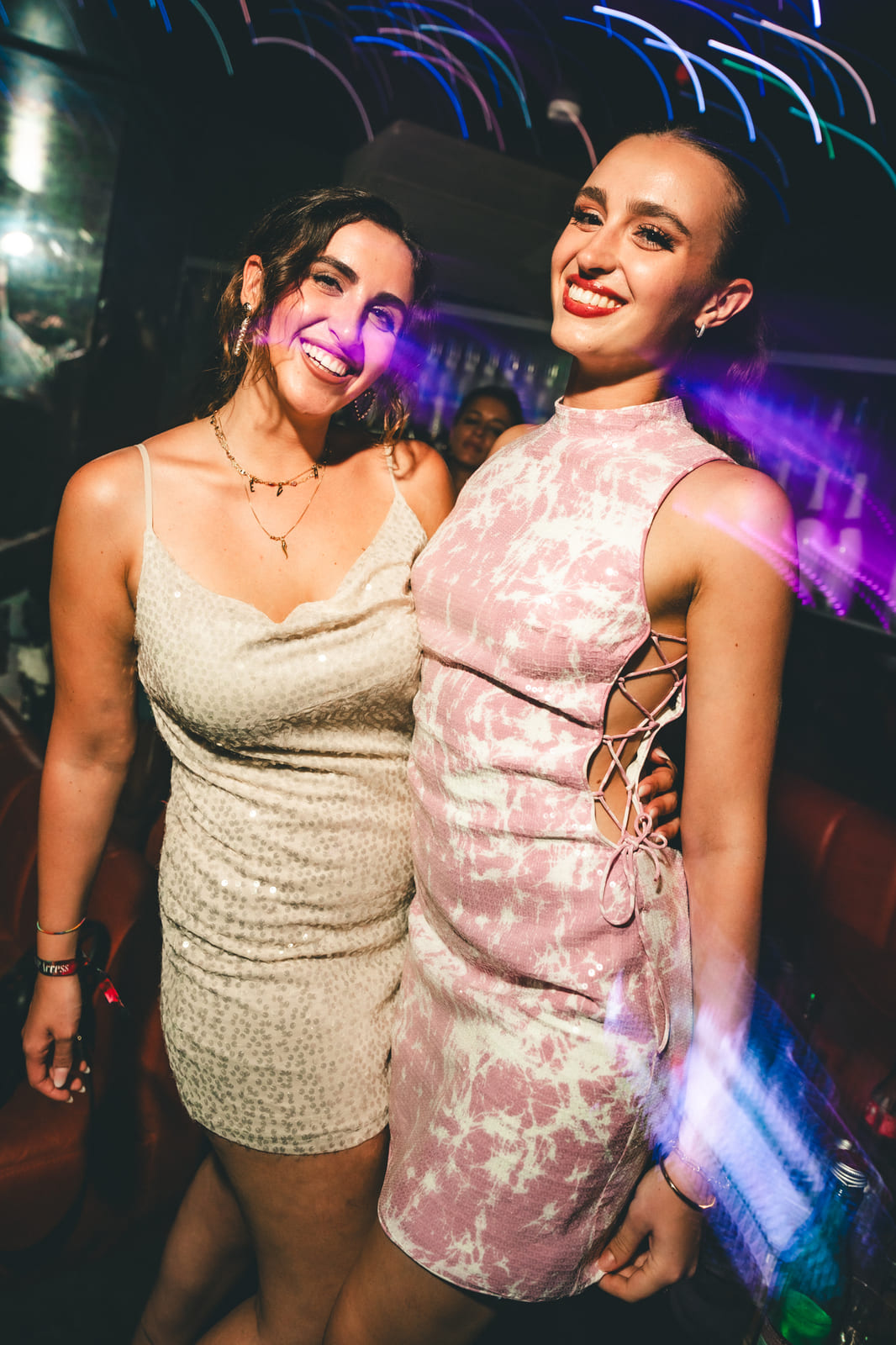 Two women smiling in sparkly dresses under colorful club lights.