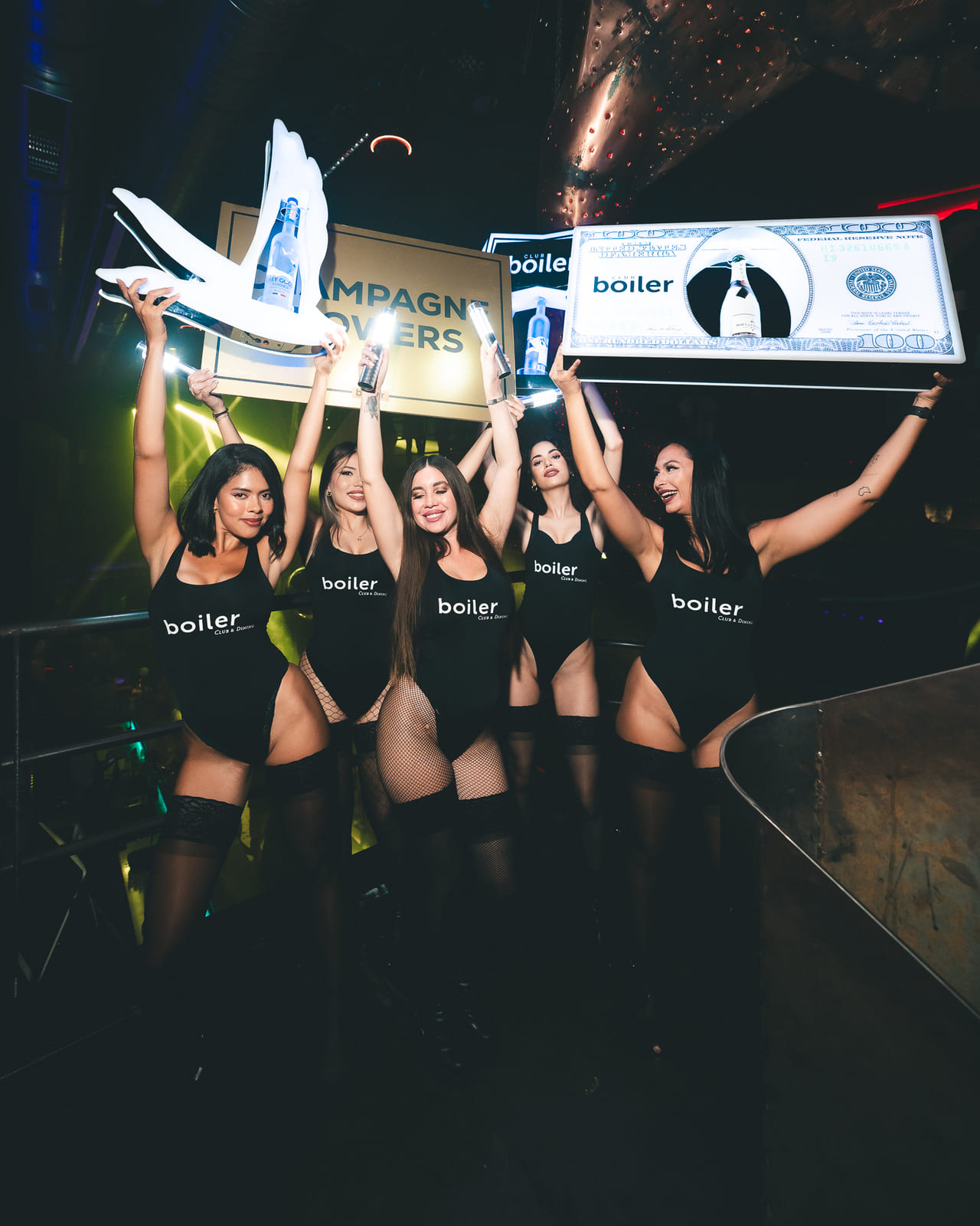 Five women in black bodysuits holding large branded signs and champagne bottles inside a nightclub.