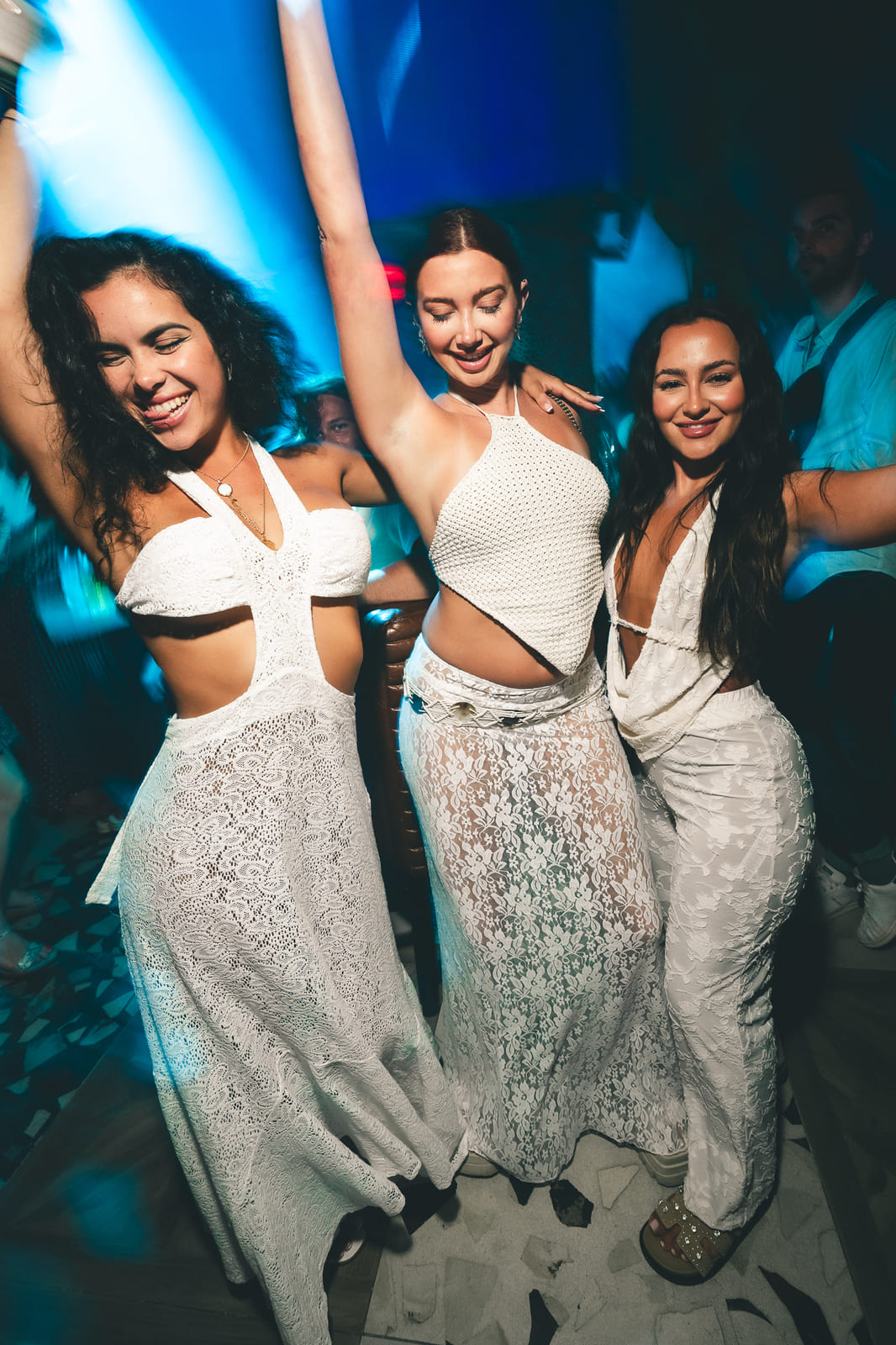 Three women in white dresses dancing joyfully under blue club lights.
