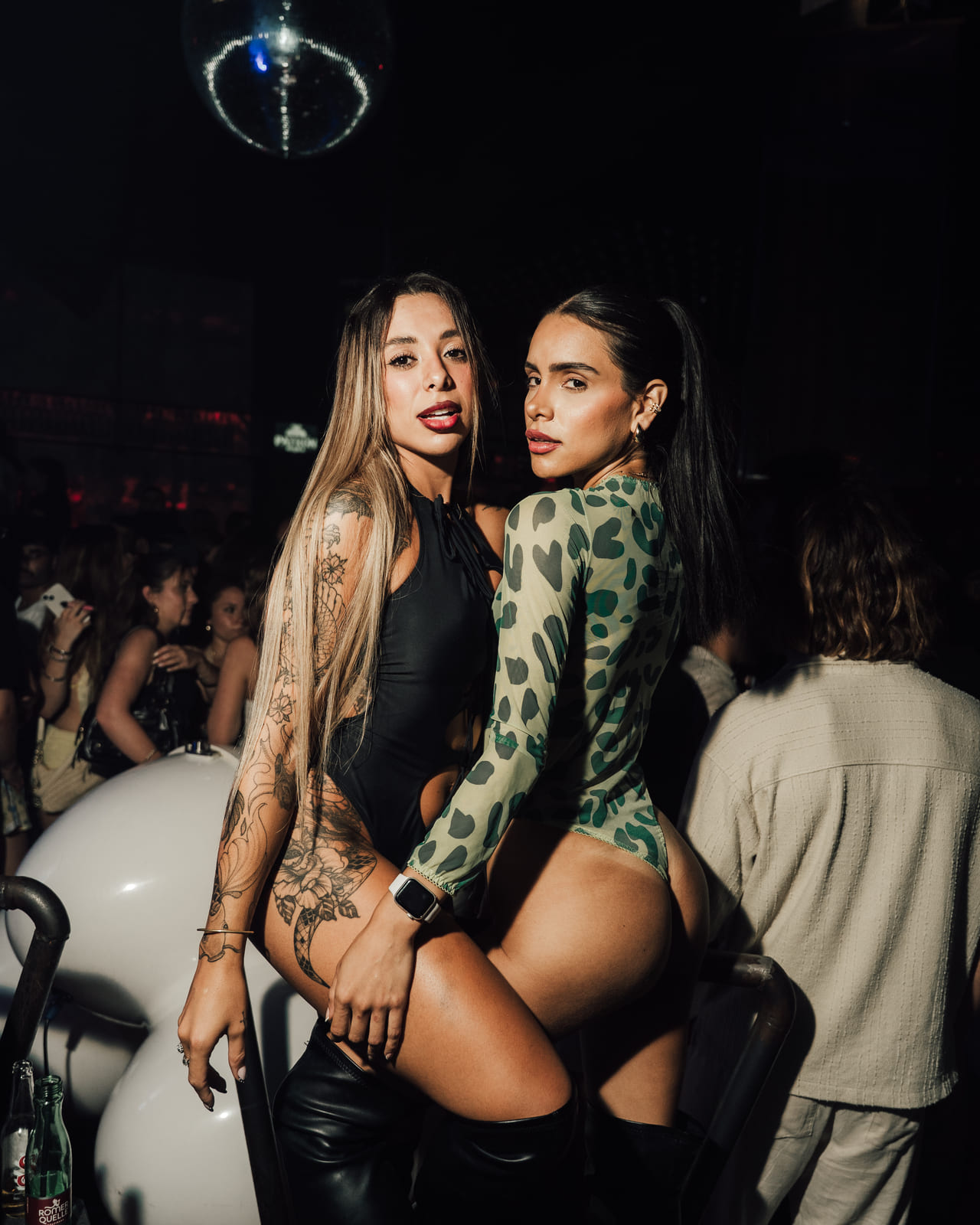 Two people posing closely on a nightclub platform, wearing stylish one‑piece outfits with a disco ball overhead.