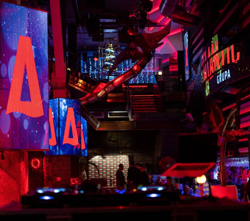 A dimly lit nightclub interior with red and blue LED screens, DJ booth in foreground, and a few people standing near the bar area in the background.