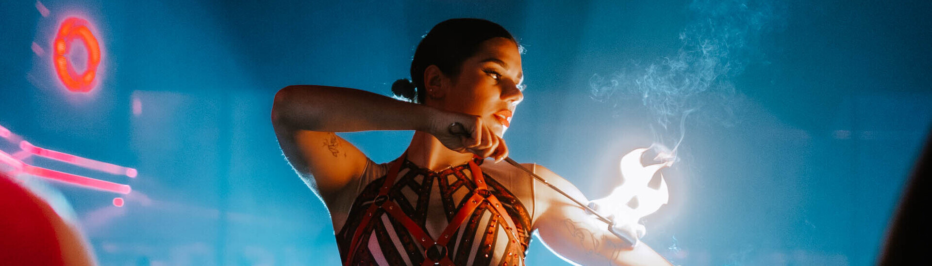 Performer in a strappy red outfit and thigh-high boots posing dramatically with fire in hand under blue and red club lighting.
