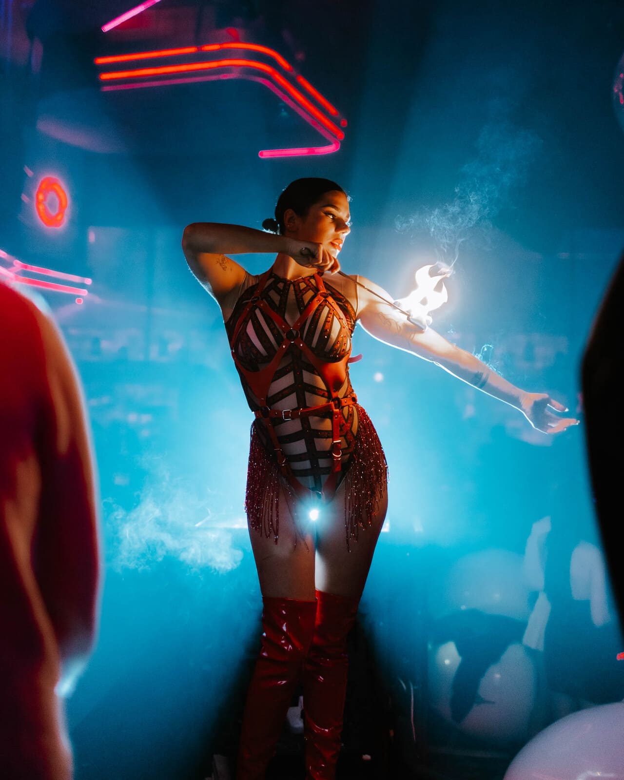 Performer in a strappy red outfit and thigh-high boots posing dramatically with fire in hand under blue and red club lighting.