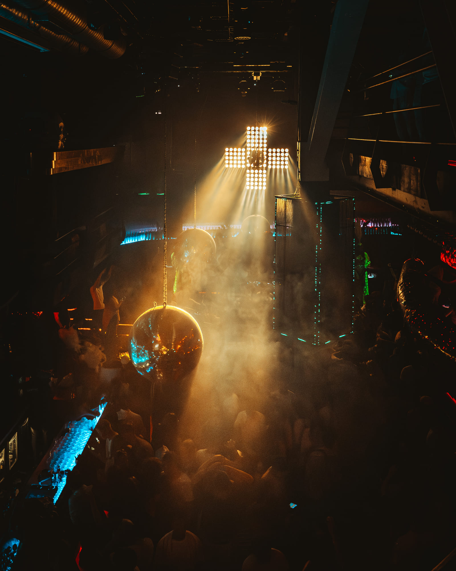 A dimly lit nightclub with a large mirrored disco ball and bright stage lighting in the shape of a cross above the dance floor. The room is filled with dense fog and a lively crowd dancing
