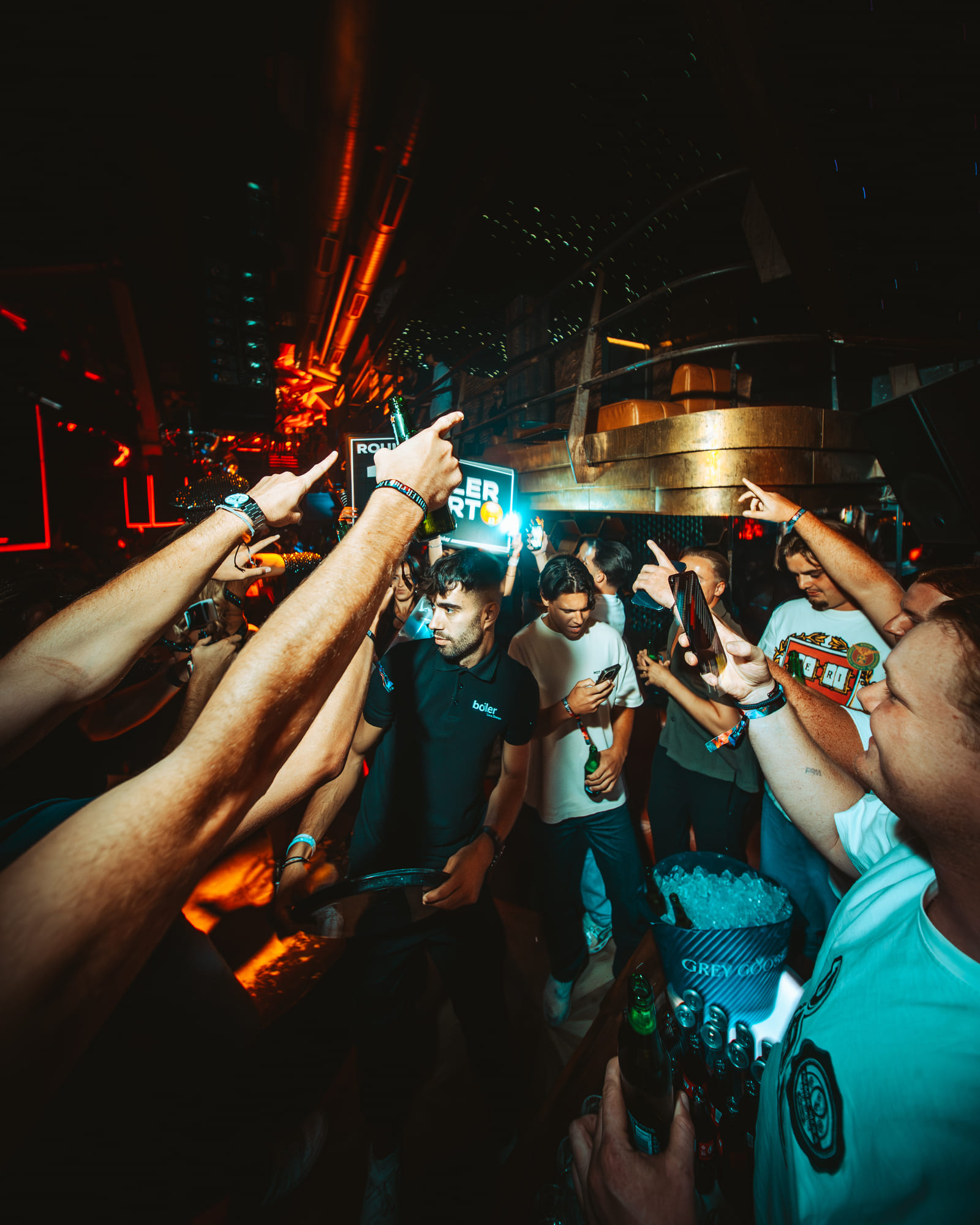 A packed nightclub scene with people raising their hands and drinks, centered on a staff member in a black "boiler" shirt holding a tray. Neon lights and bottle service are visible.