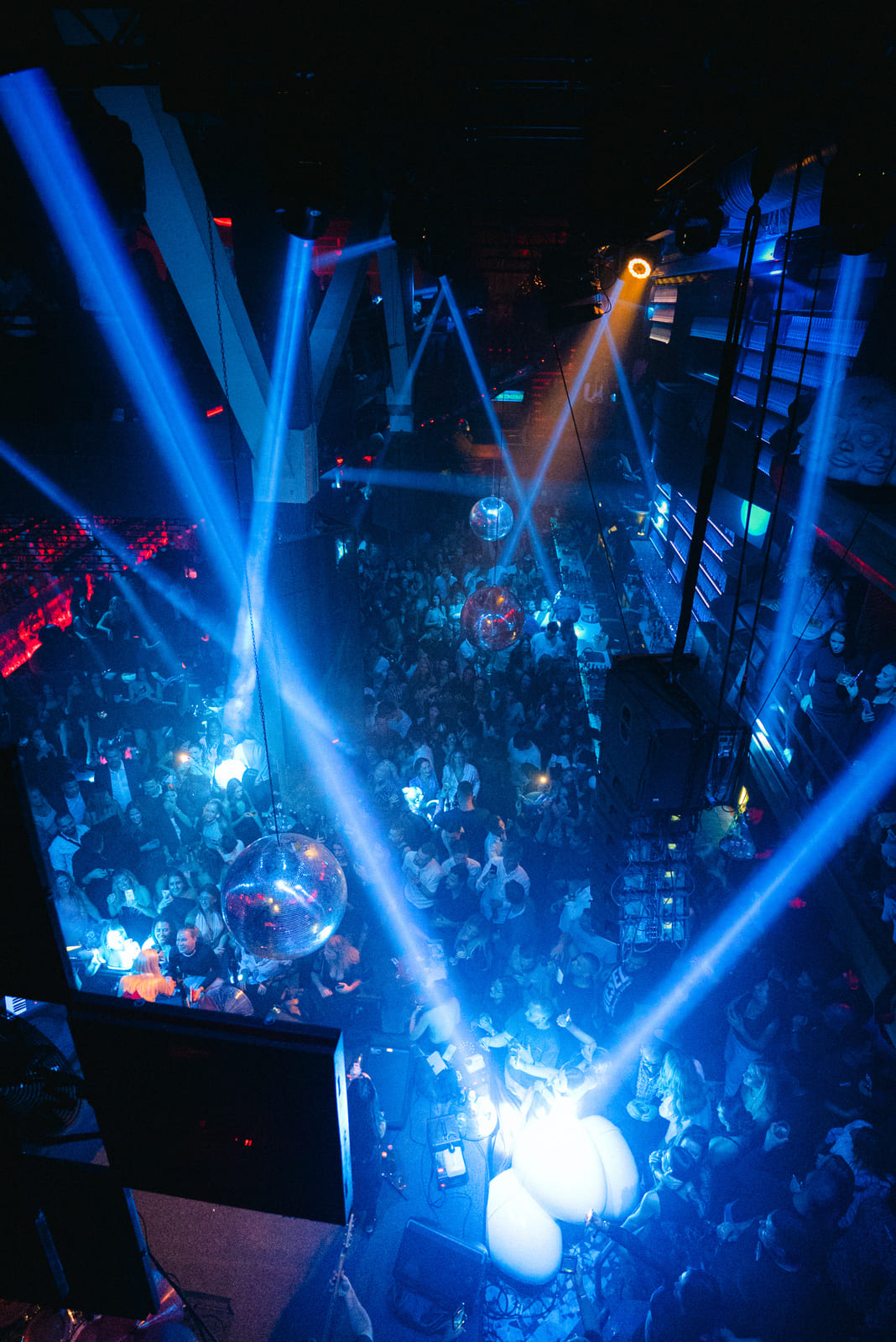 Overhead view of a crowded nightclub dance floor with bright blue spotlights and hanging disco balls.