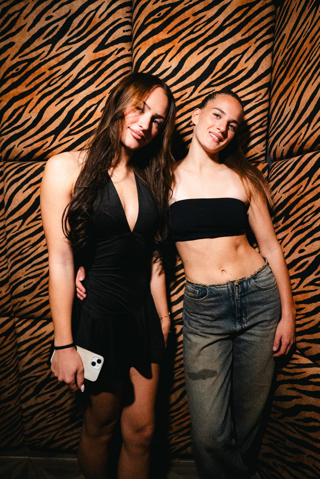 Two friends posing together in front of a tiger‑print wall, one wearing a black dress and the other jeans and a black top.