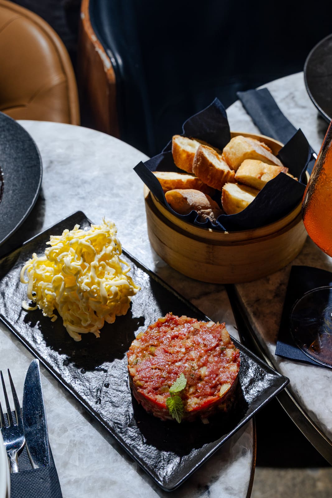 Plate with beef tartare, shredded cheese, and a bamboo basket of sliced baguette on a marble table.
