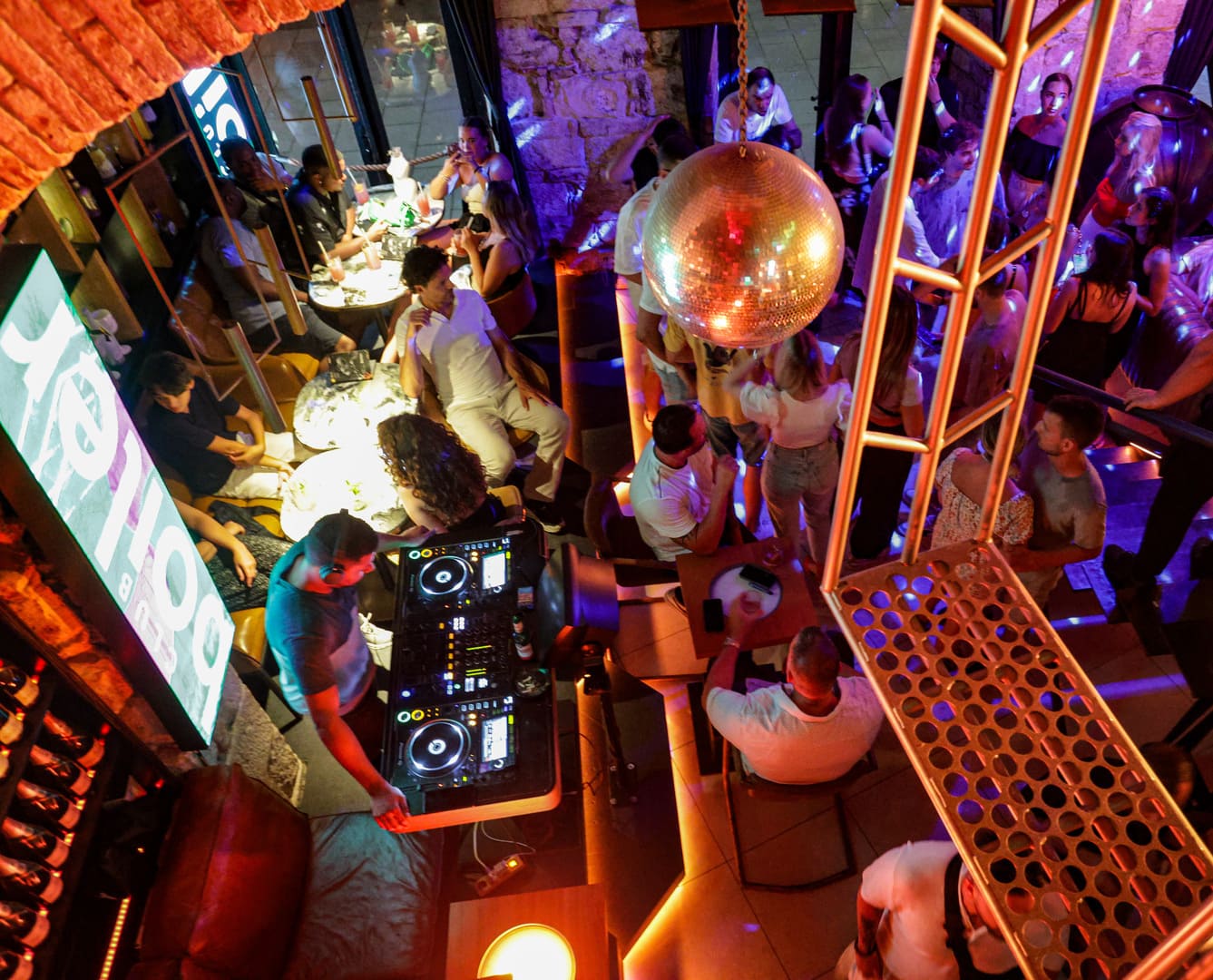 Overhead view of a lively nightclub scene with a DJ performing, people dancing under a disco ball, and groups seated at tables enjoying drinks and conversation.
