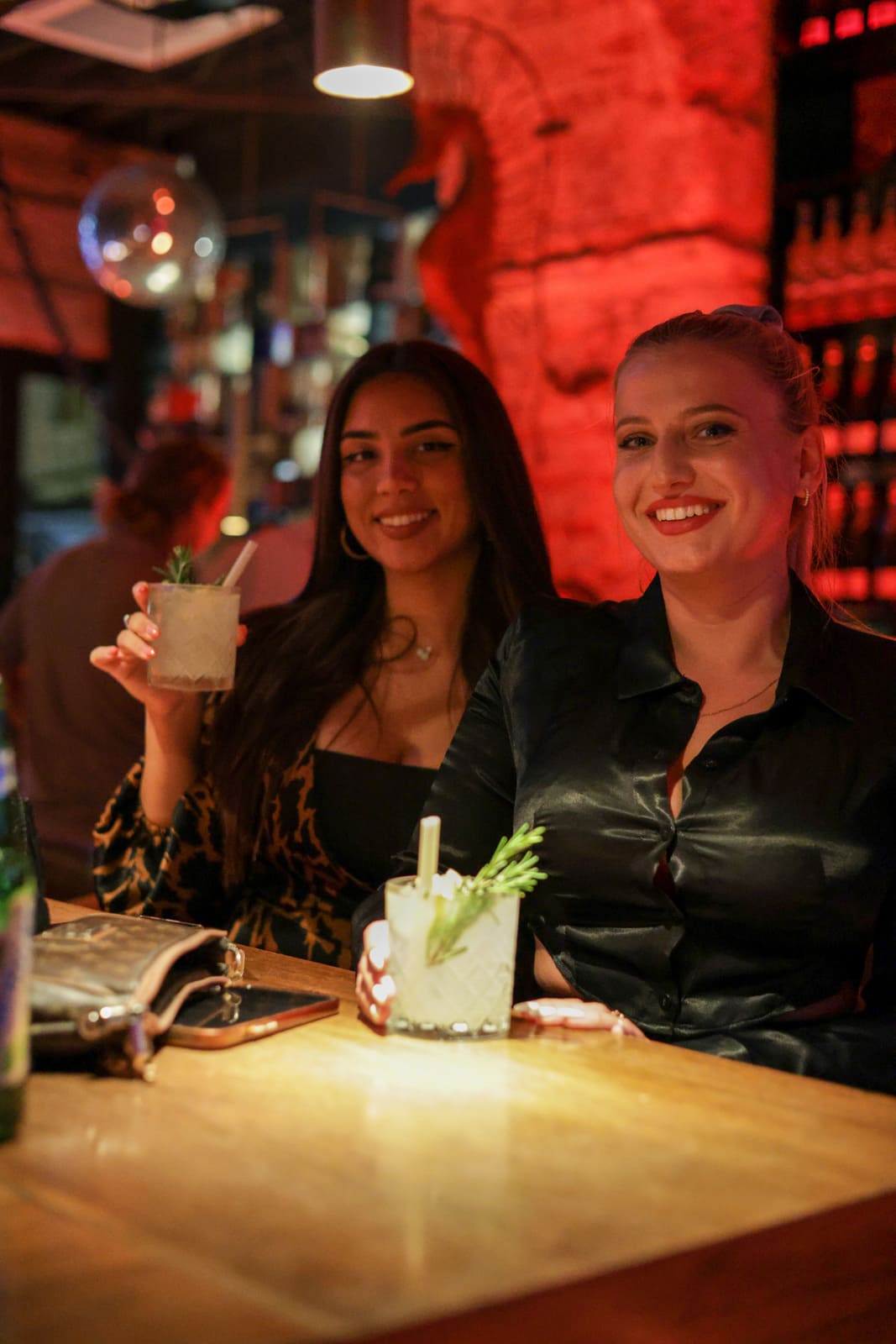 Two women sitting at a bar holding cocktails, smiling at the camera.