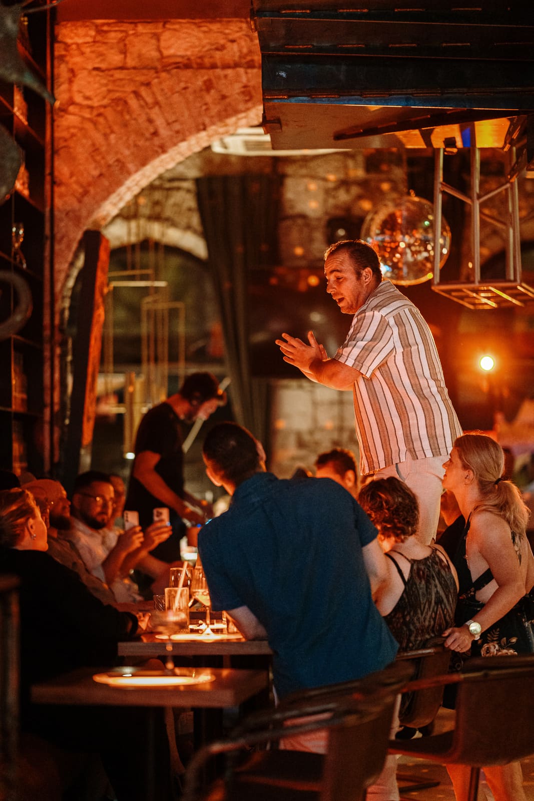Man standing on a bench speaking to a group inside a stone-walled restaurant