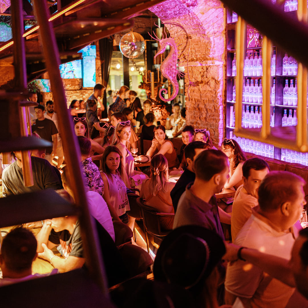 Crowded indoor bar scene with groups of people seated and standing under warm pink and purple lighting, with a glowing seahorse decoration and bottles lit on a backlit shelf.