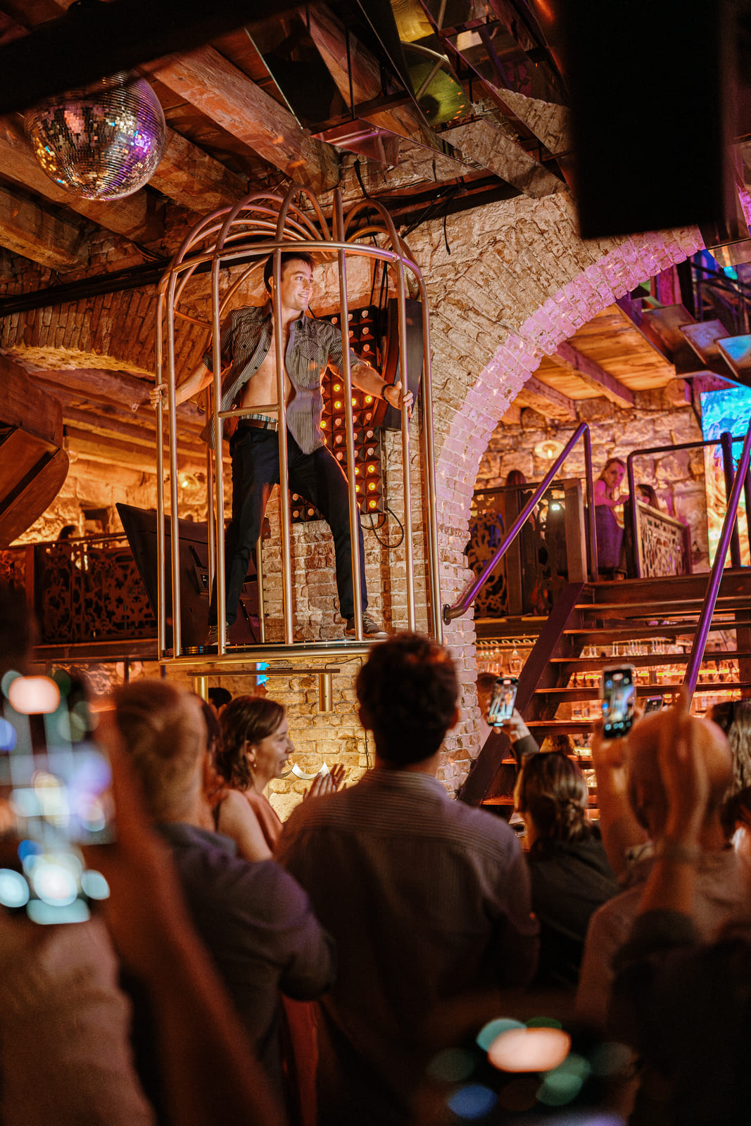 Shirtless man dancing inside a gold cage as crowd records with phones
