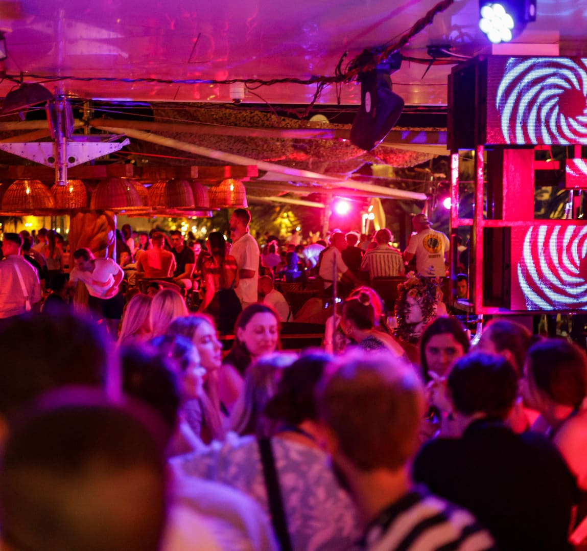 A packed nightclub with vibrant red and purple lights illuminating a diverse crowd of guests enjoying drinks and music under a covered patio.