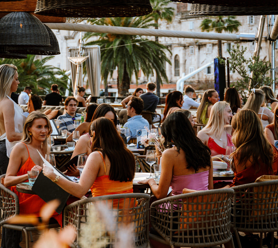 Groups of stylishly dressed people enjoying food and drinks on a lively rooftop patio with palm trees and historic buildings in the background.