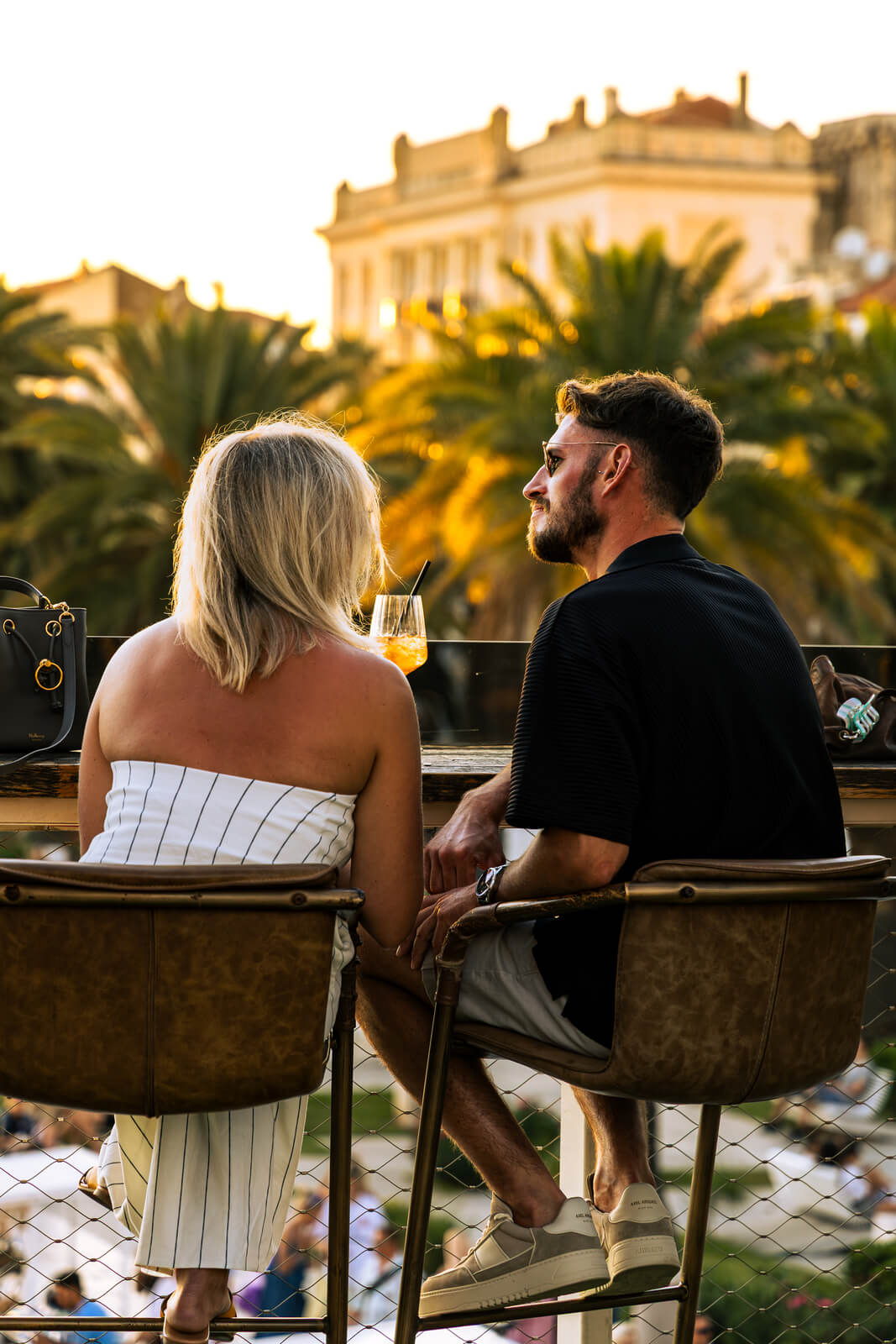 Couple sitting at an outdoor rooftop bar at sunset with drinks and palm trees in the background.