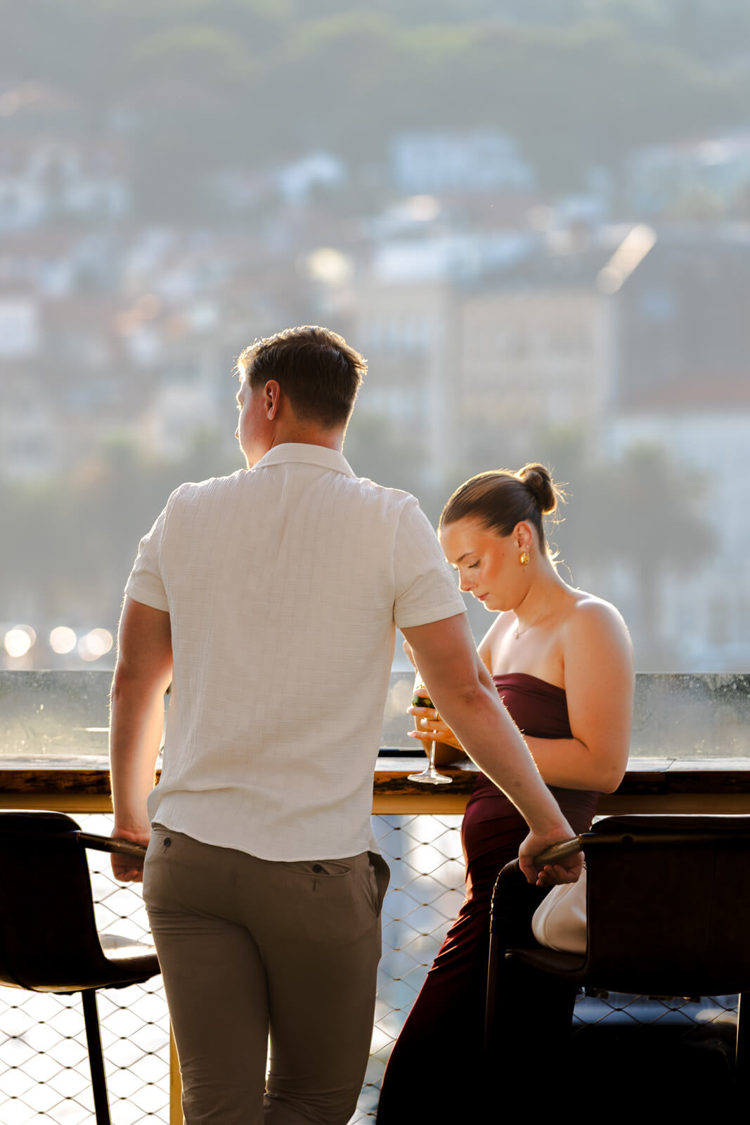 Couple enjoying drinks on an outdoor terrace at sunset, overlooking a cityscape.