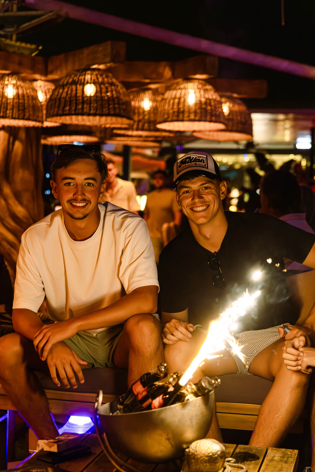 Two friends sitting at a lounge table at night with a sparkler and bottle service in front of them.