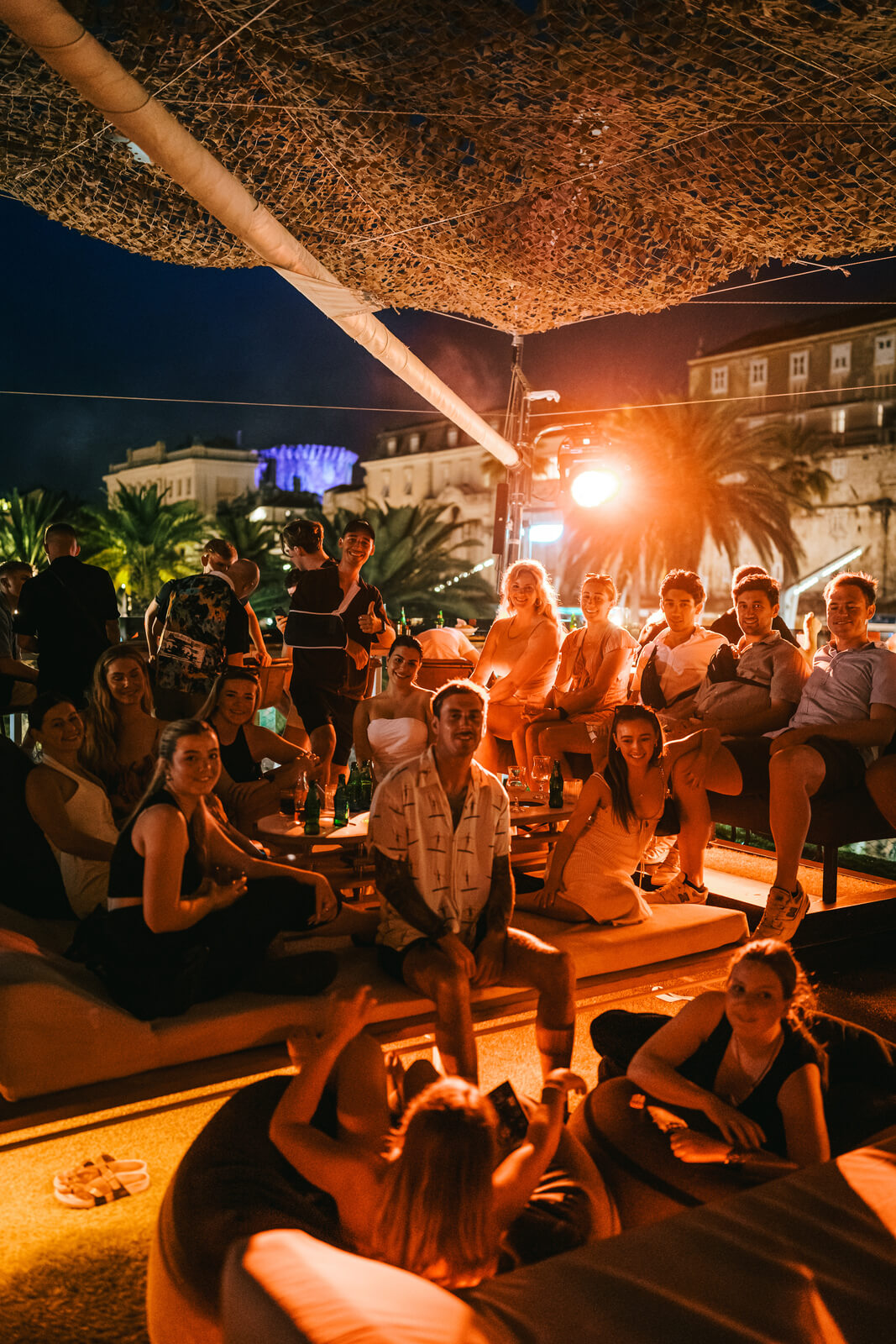A lively group of friends sitting on large bean bags and cushions on a crowded rooftop terrace at night, illuminated by a warm, overhead light source.