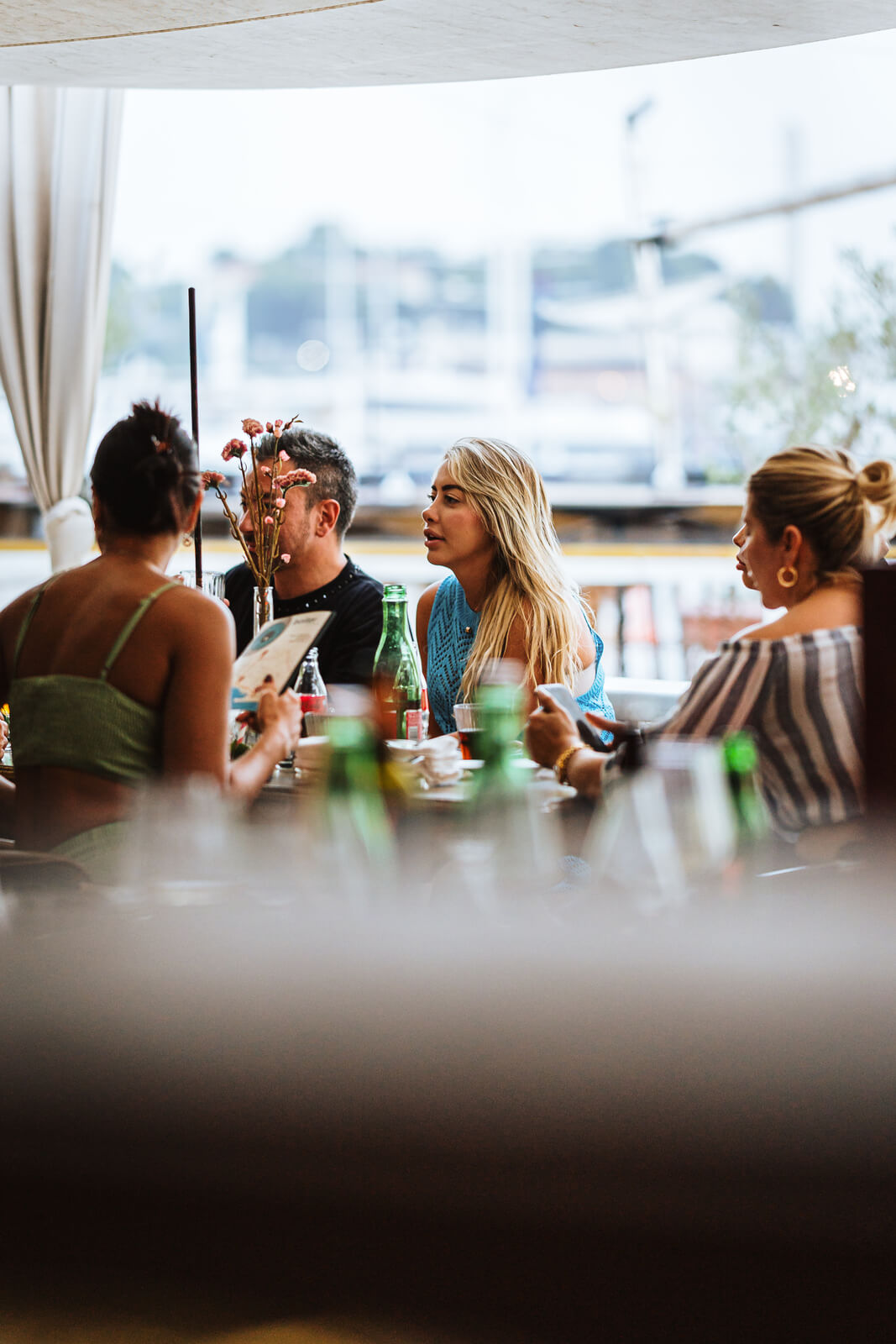 Four friends seated around a table at an outdoor cafe with a bright background view of a harbor. The focus is on a blonde woman in a blue top talking to the man across from her.