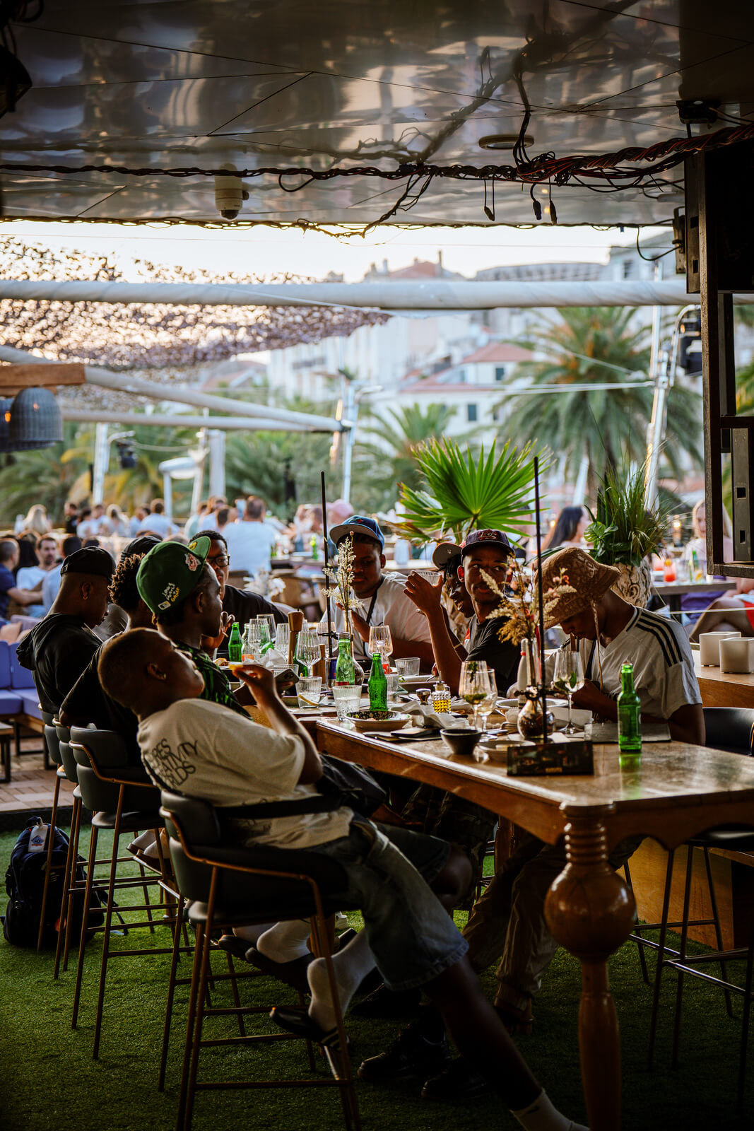 Group of men sitting at a long outdoor table sharing food and drinks under soft evening light.