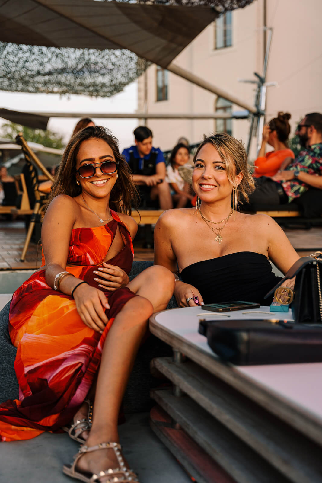 Two smiling women sitting on an outdoor lounge at sunset, dressed in elegant evening attire.