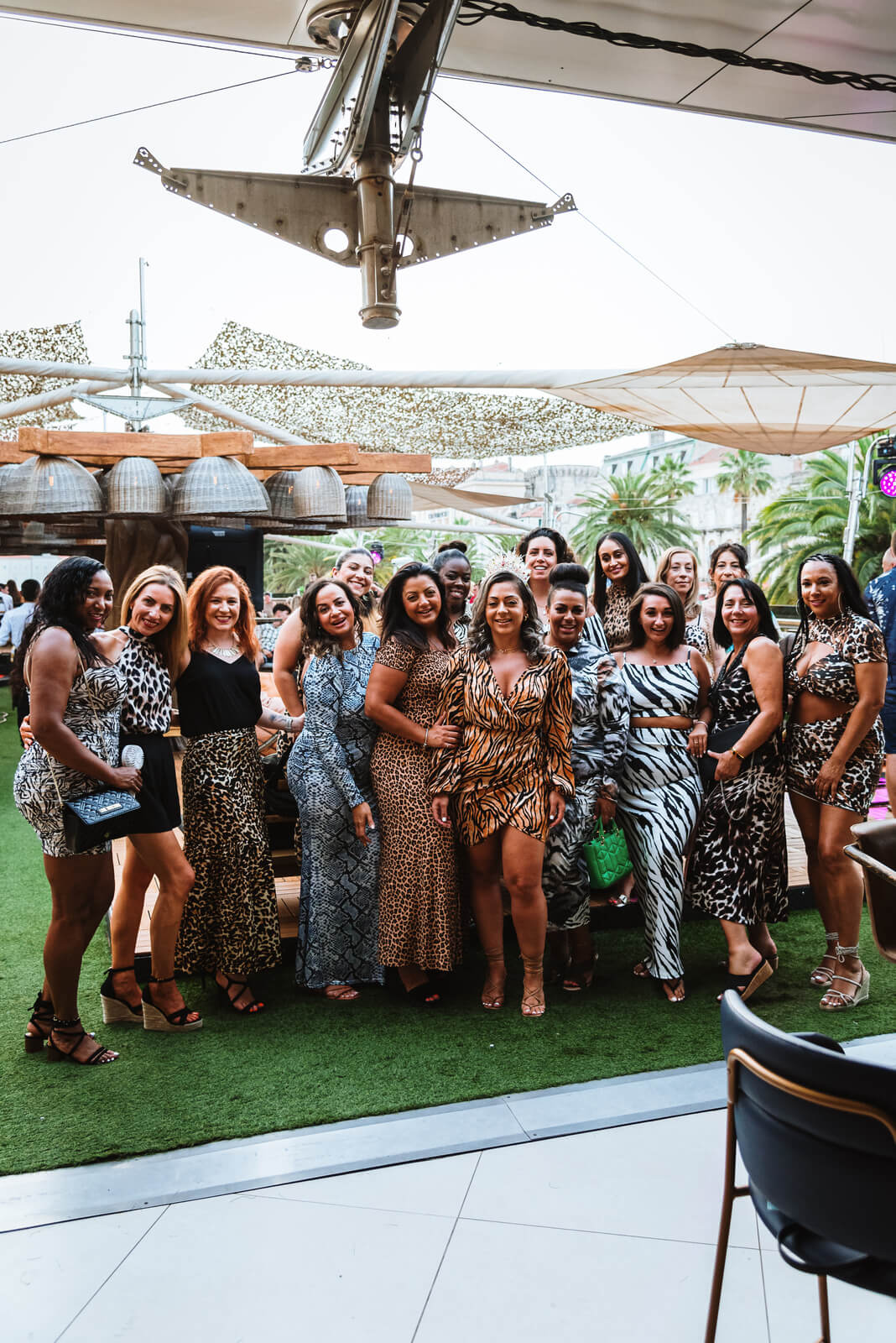 A large group of diverse women posing together for a photo in a group hug on a rooftop terrace. Most of the women are wearing various animal print dresses and outfits.