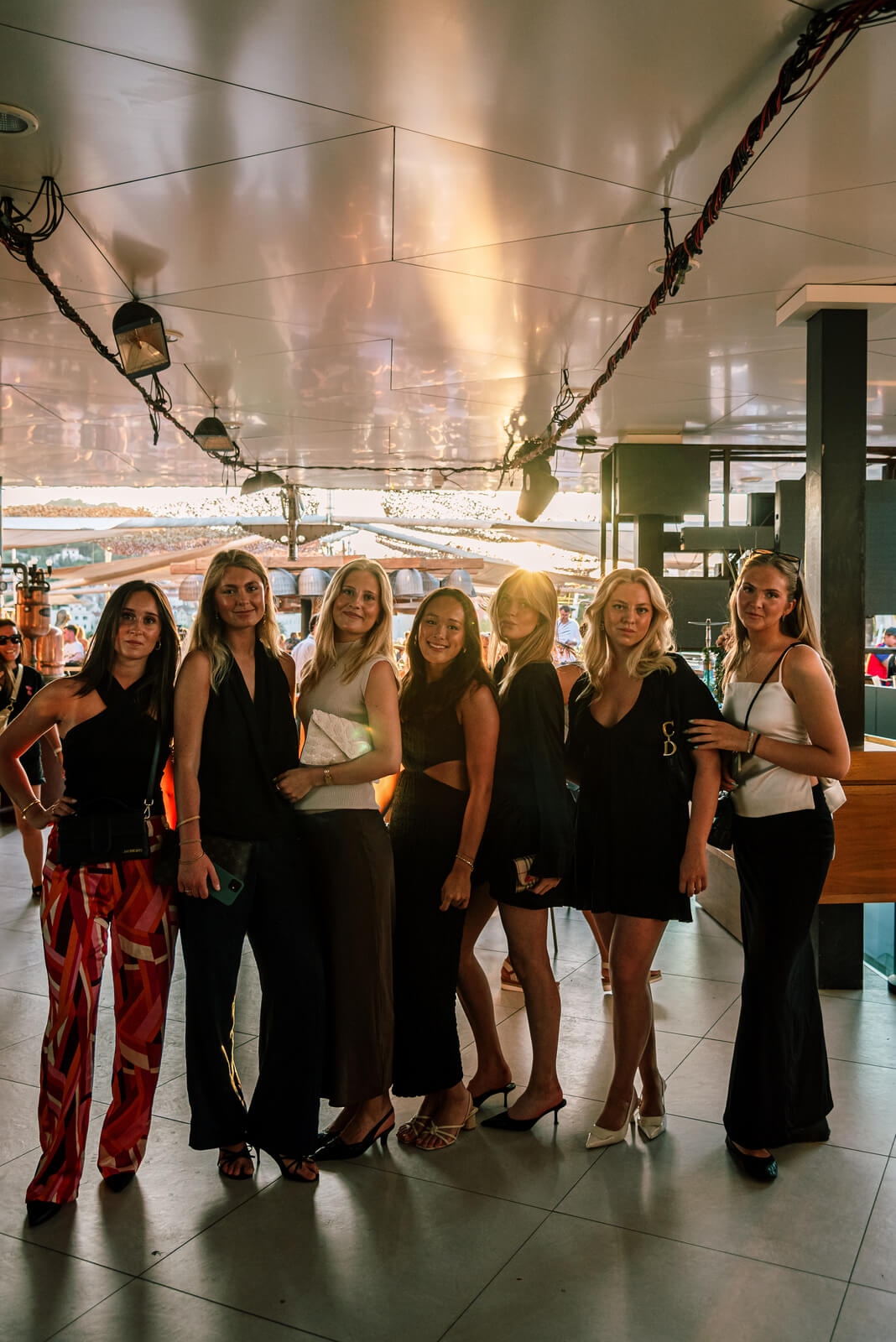 A group of six fashionable women posing elegantly side-by-side in dark evening wear on a bright indoor/outdoor deck under a string of lights.