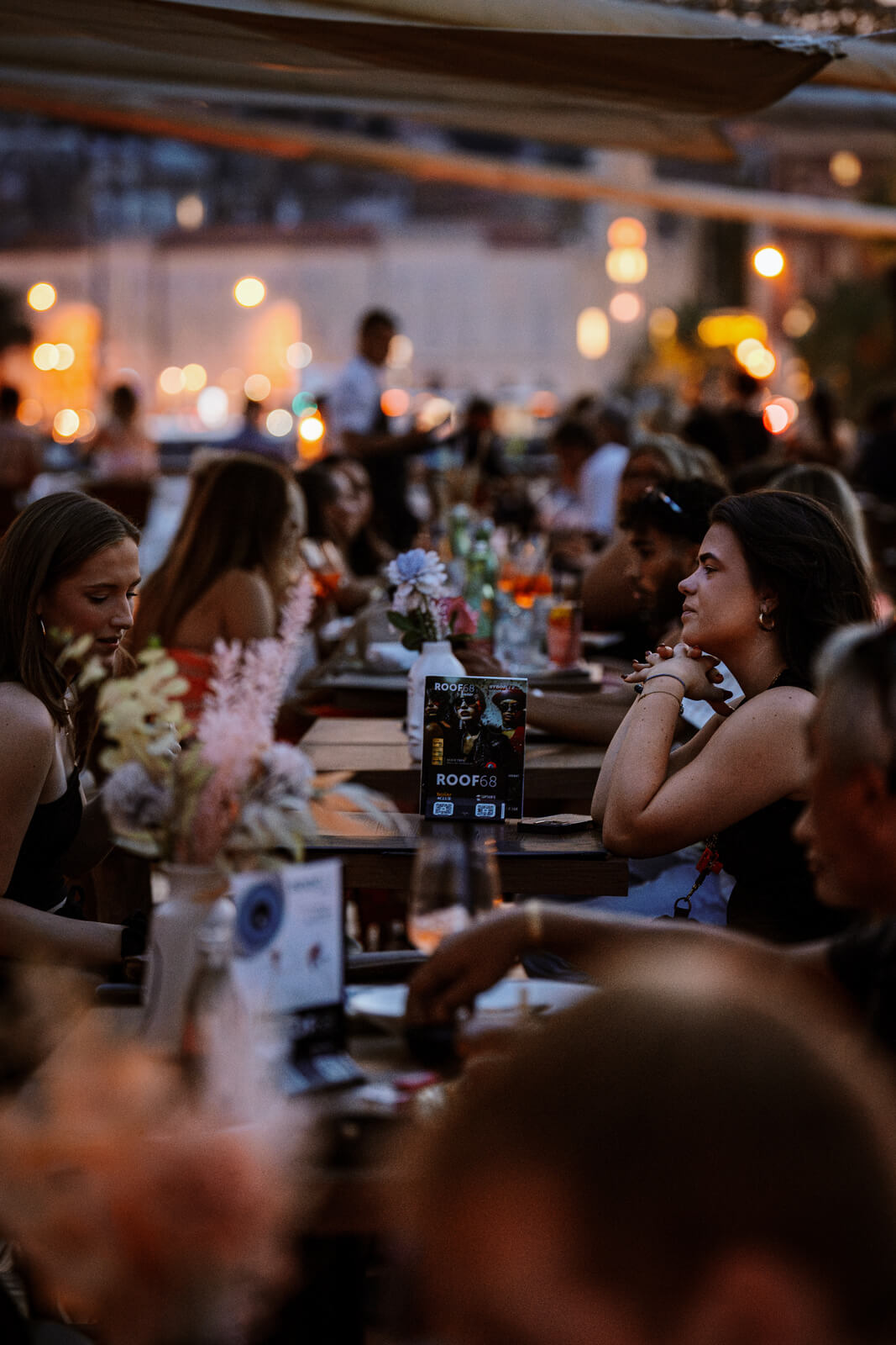 Guests seated at wooden tables outdoors during twilight, enjoying drinks under canopy lights.