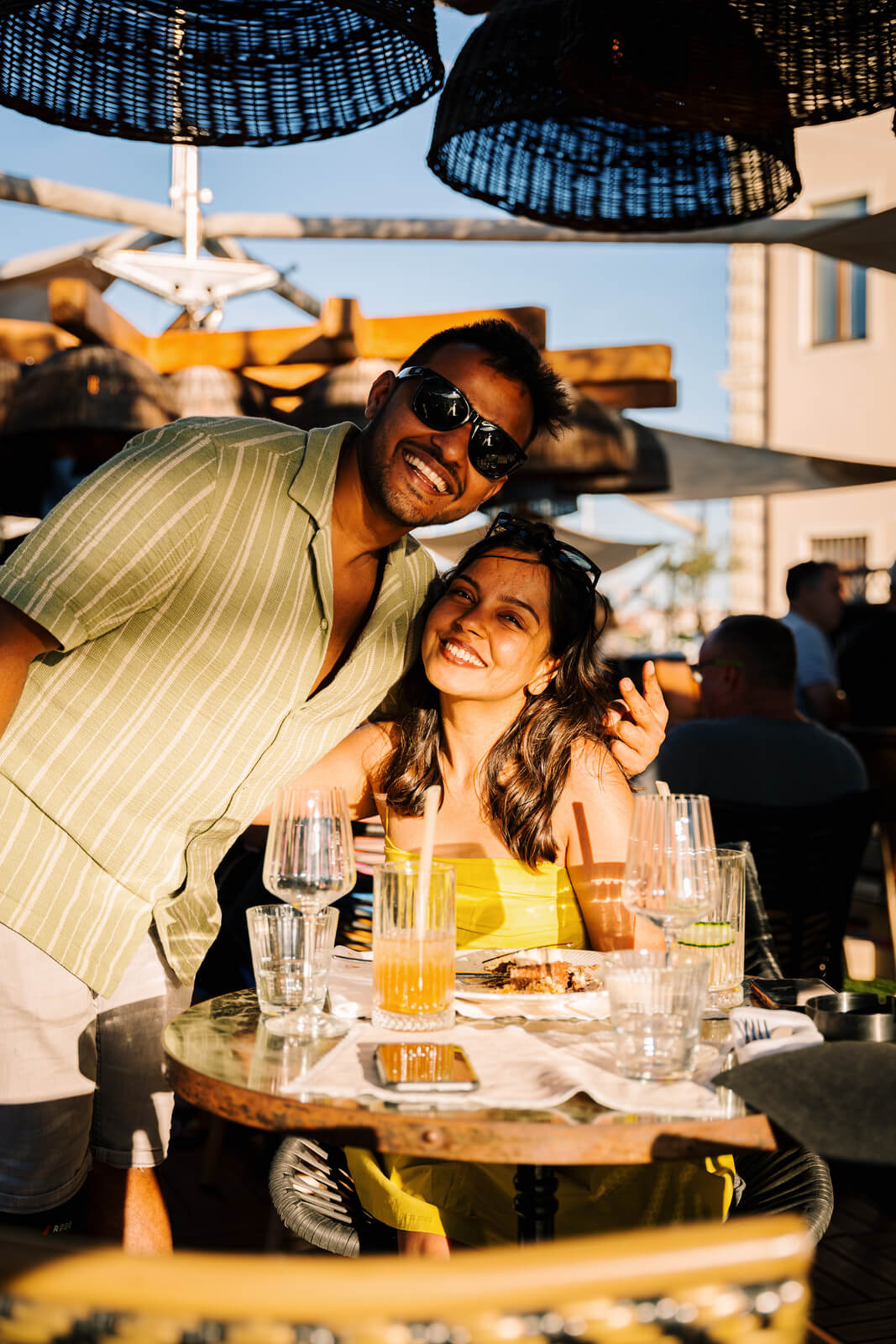 A smiling young couple hugging and posing for a close-up photo while seated at an outdoor restaurant table during golden hour. The table is set with drinks and plates.