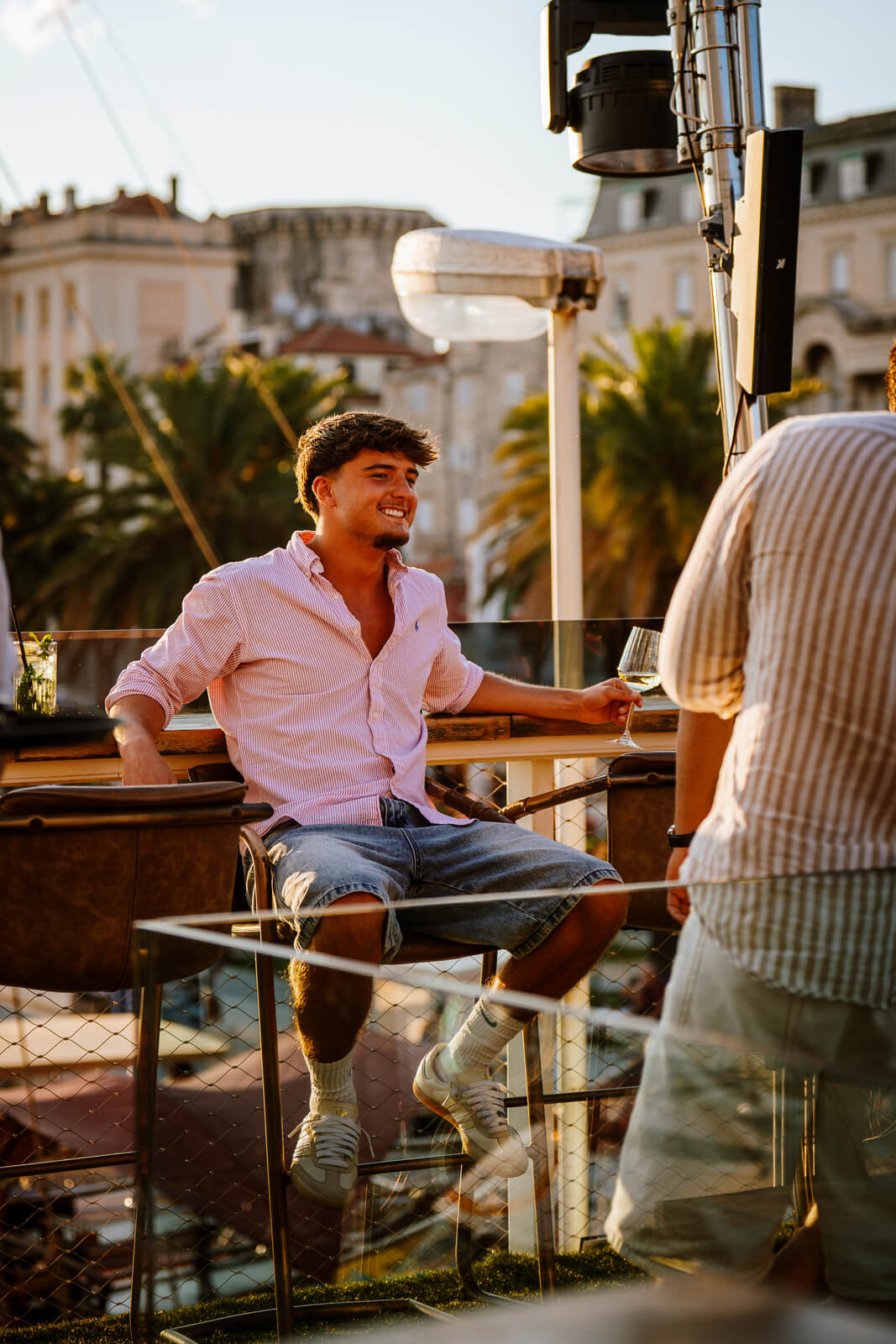 Man in pink shirt smiling and holding a drink at a rooftop bar during sunset with palm trees behind him.