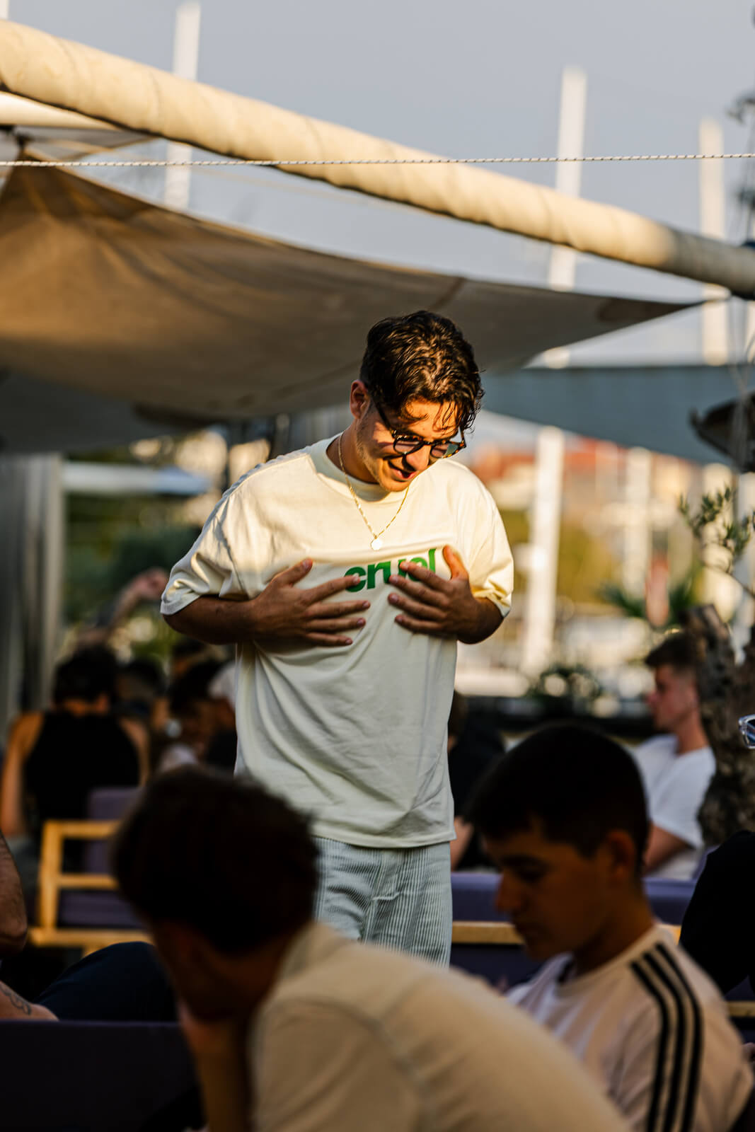 Man smiling while standing outdoors at a café, surrounded by people in casual summer clothing.