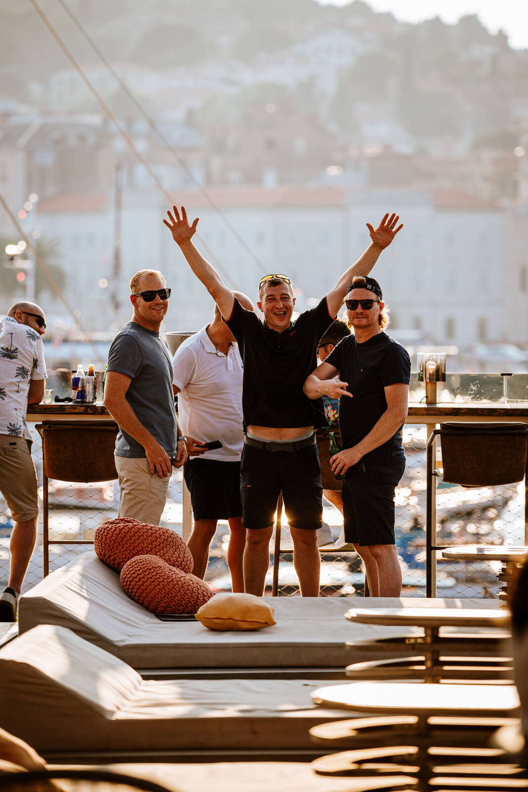 A group of four smiling men standing outdoors on a bright rooftop deck, with the central man raising his hands in excitement. They are casually dressed, enjoying the sunny day with a city and harbor view in the blurred background.
